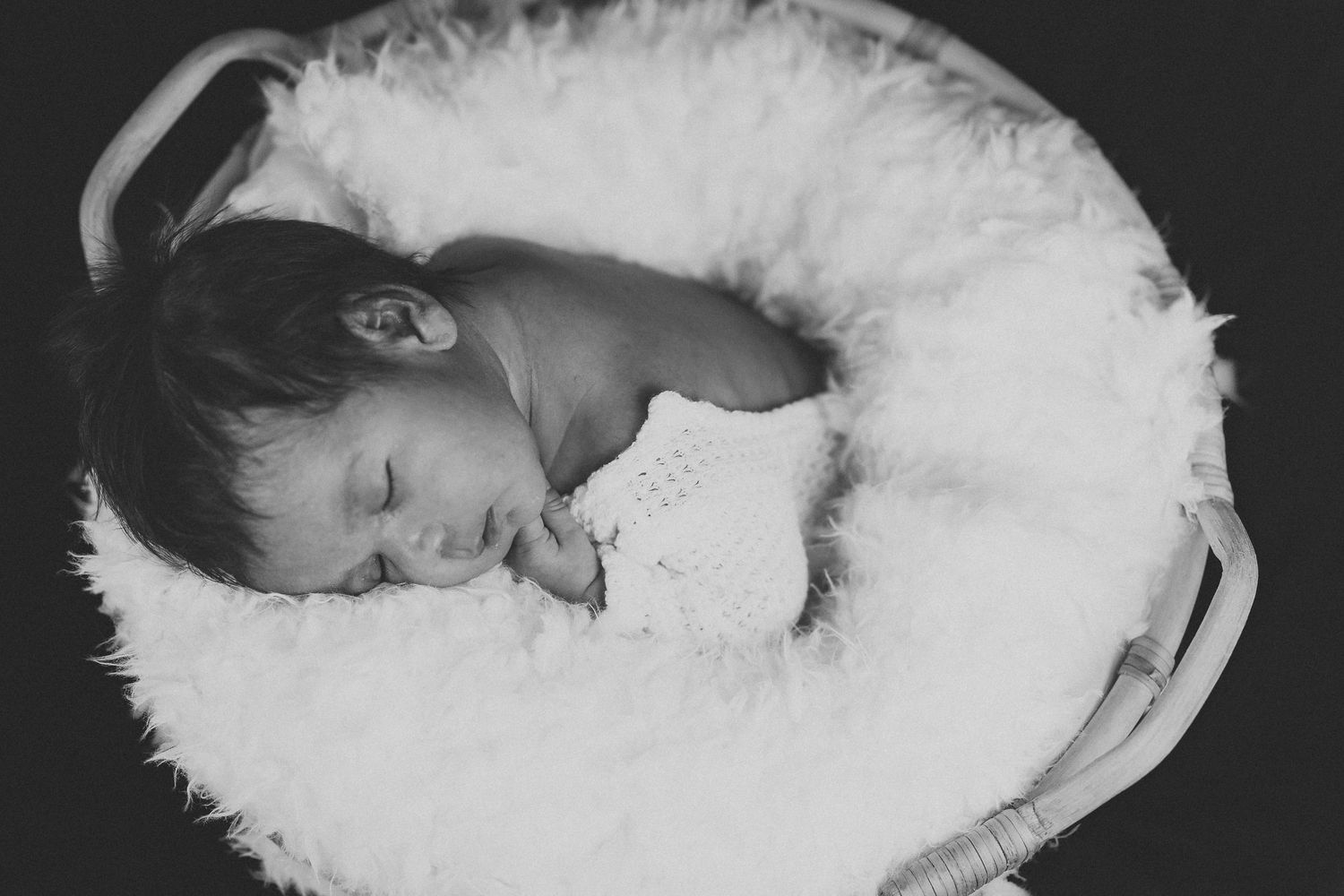 Newborn baby rests peacefully on white fluffy blanket in black and white portrait.