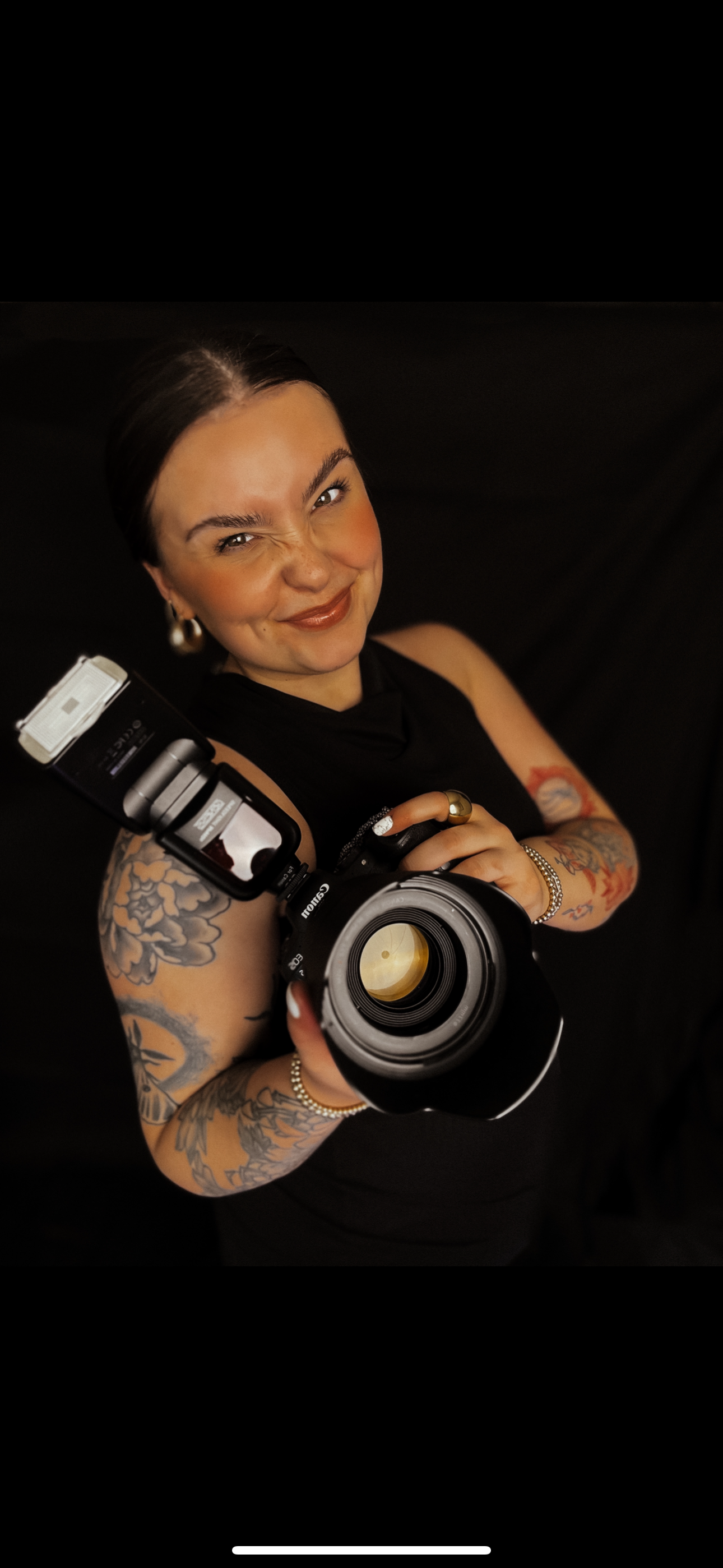A photographer with tattoos holding a professional camera with flash against a black background.