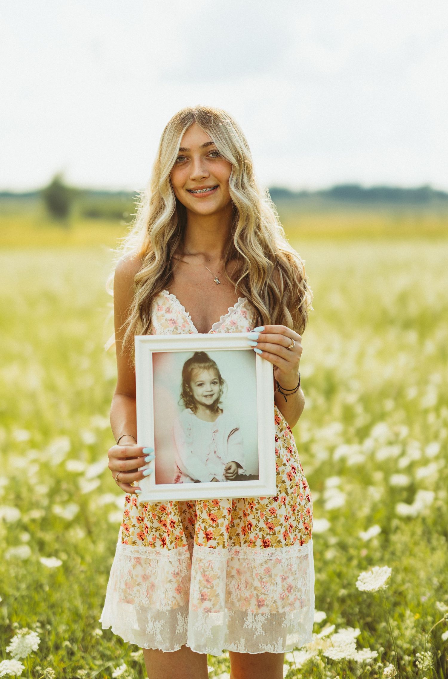 Someone in a floral dress holding a framed childhood photograph in a daisy field.