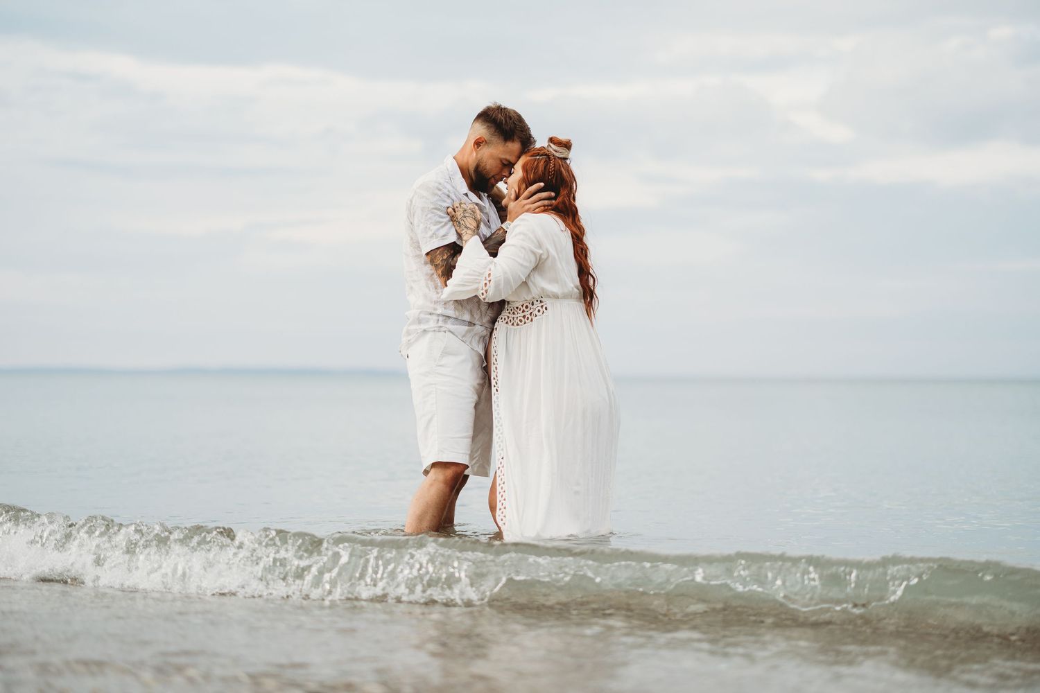 Couple embracing in shallow water wearing white outfits.