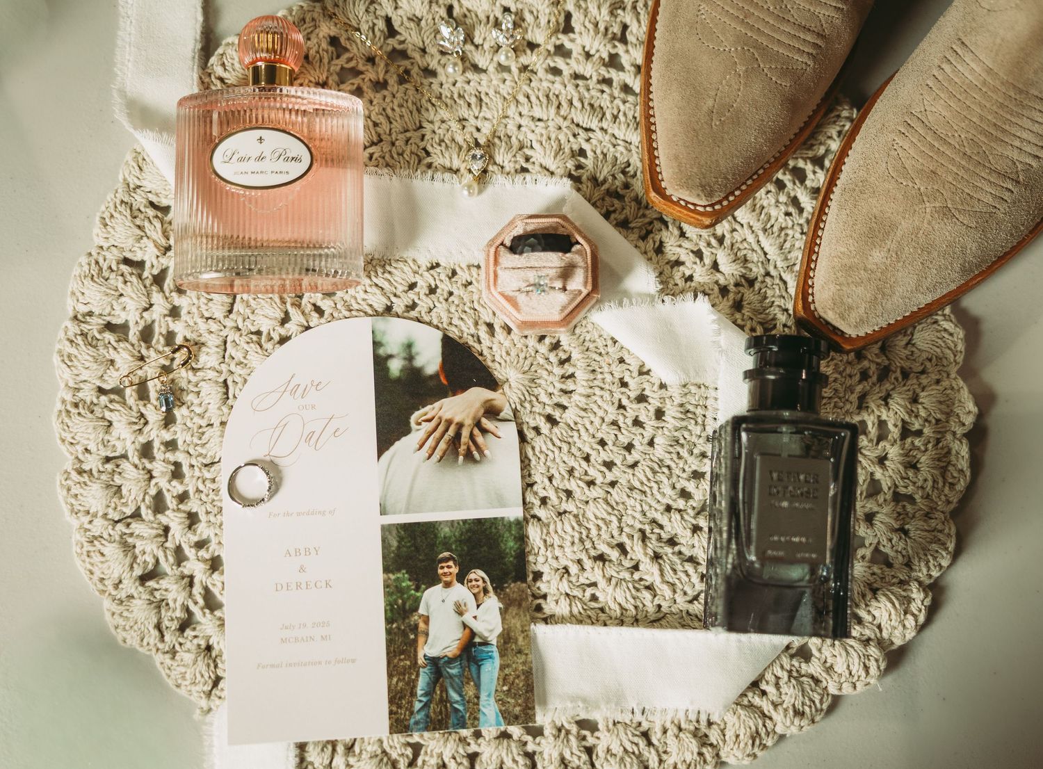 Wedding details flatlay with perfume bottles, rings, and photographs arranged on vintage lace doily.