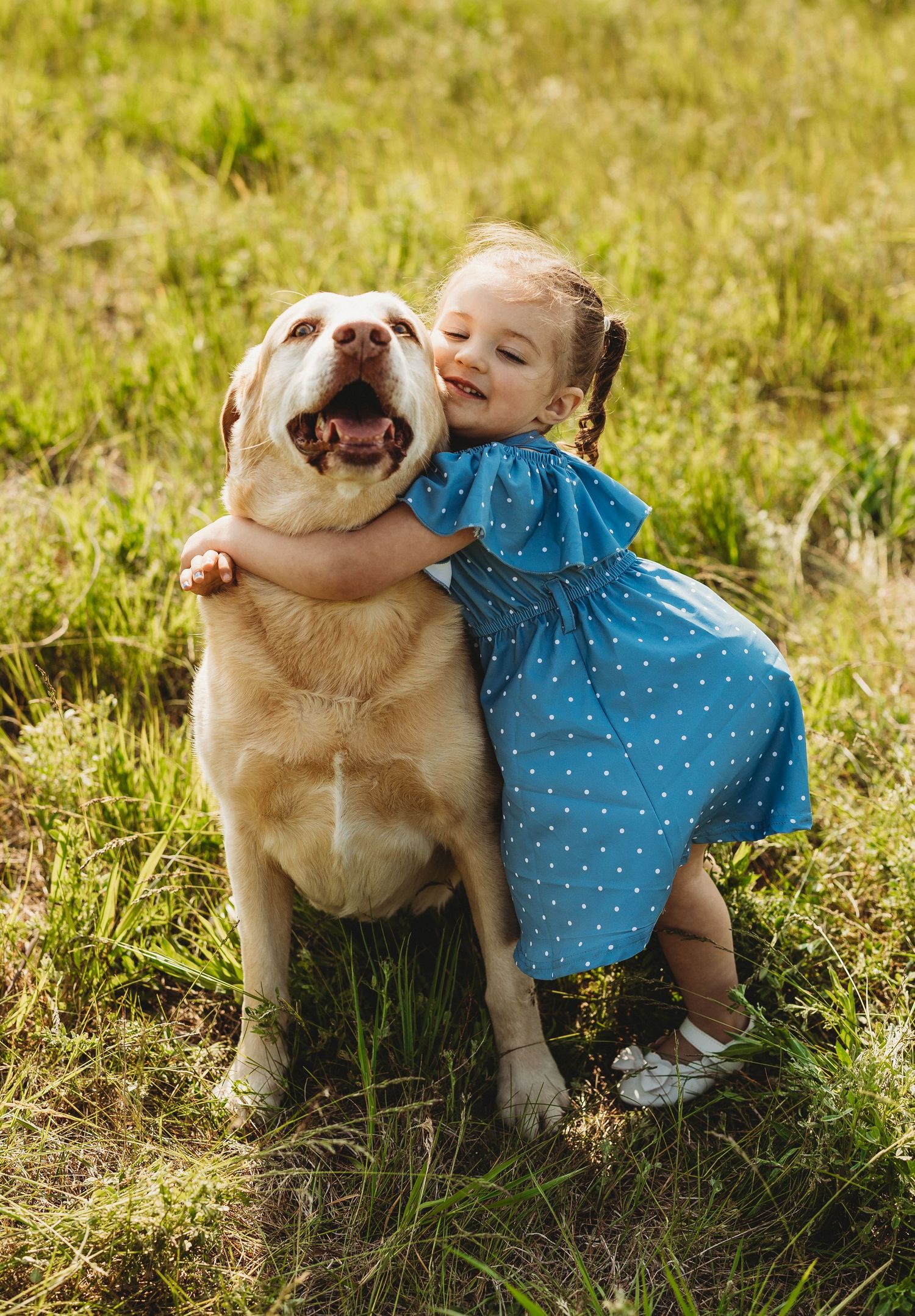 A child in a polka dot dress plays with a friendly golden retriever dog in the sunlit grass.