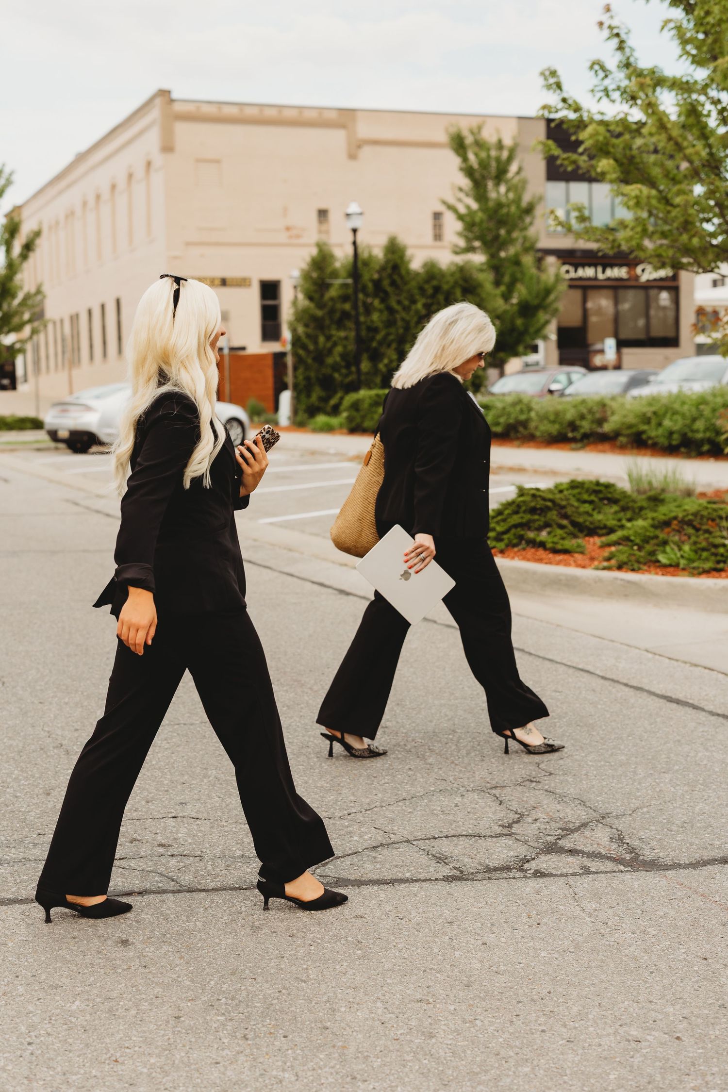 Two women in matching black jumpsuits and heels walking across a parking lot, one holding a phone and the other carrying a bag.