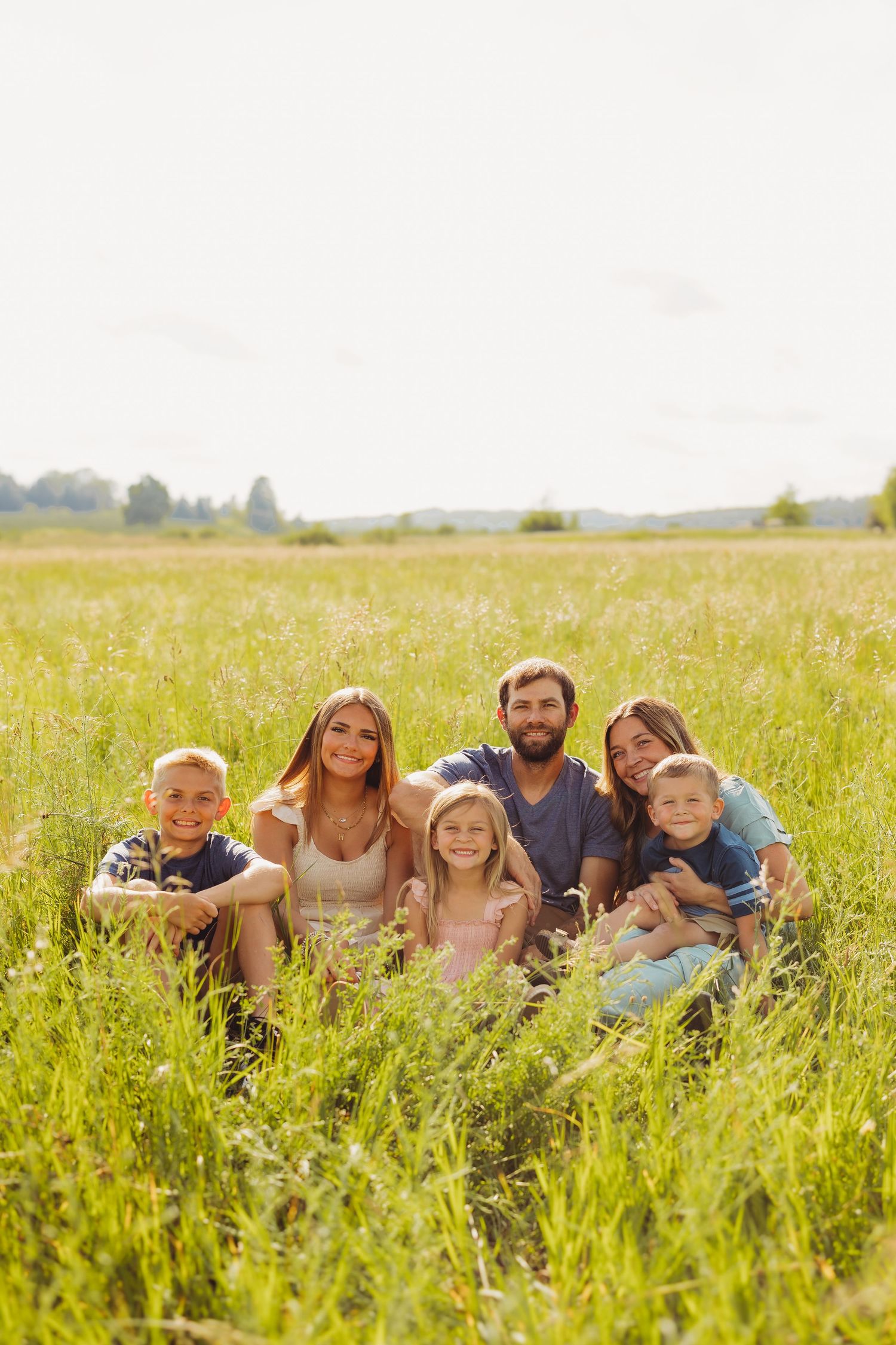 Happy family of five sitting together in a sunny green meadow, smiling at the camera on a bright summer day.
