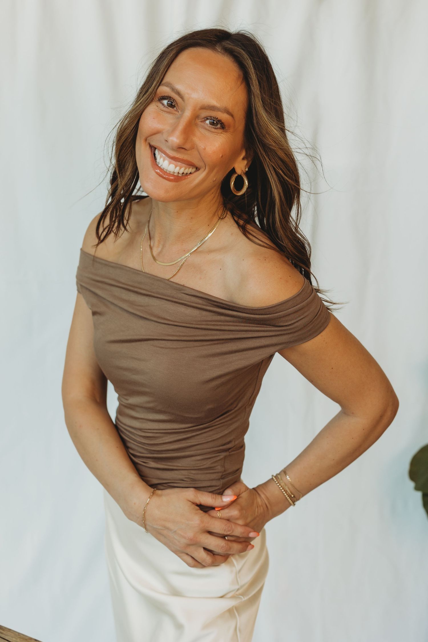 Woman smiling in brown off-shoulder top, white skirt, gold hoop earrings and bracelet, posing against white backdrop.