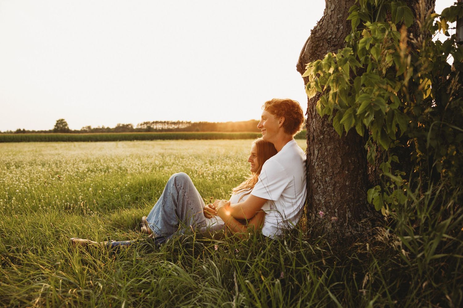 Couple sitting together against a tree in a golden sunset field, enjoying a peaceful romantic outdoor moment.