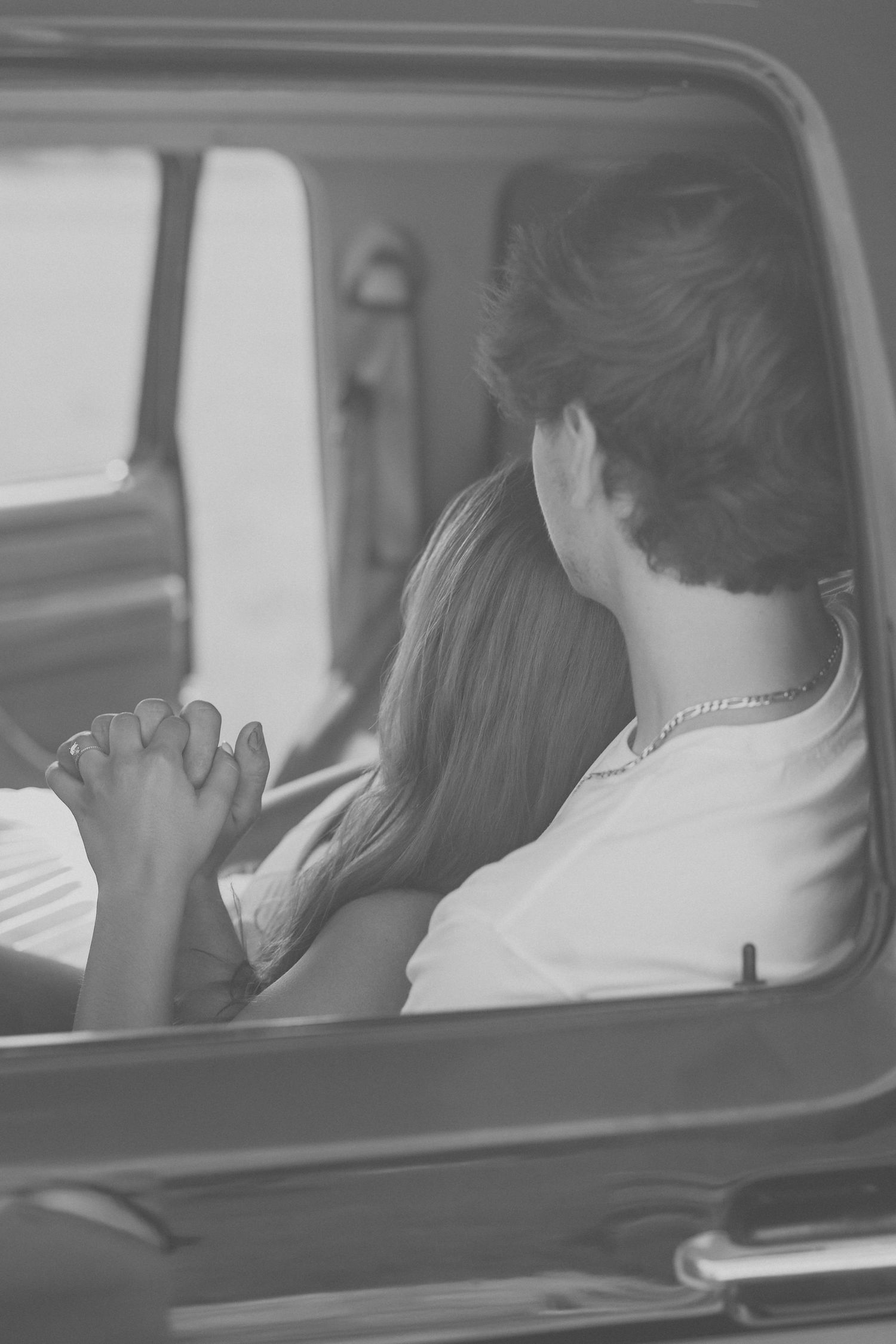 Black and white photo of a couple holding hands inside a vintage vehicle, viewed from behind.