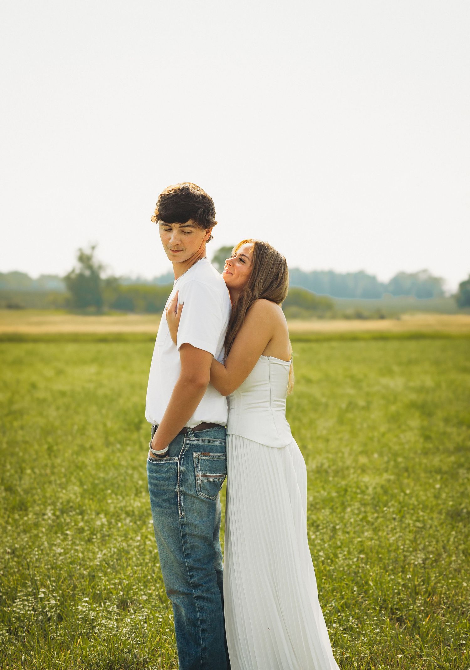 A young couple poses in a green field, the girl in white hugging the boy from behind as golden sunlight glows around them.