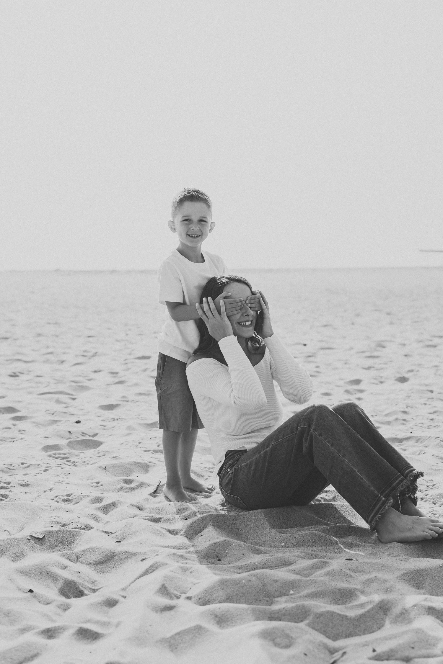 Black and white photo of a boy and woman laughing together on a sandy beach near the ocean.