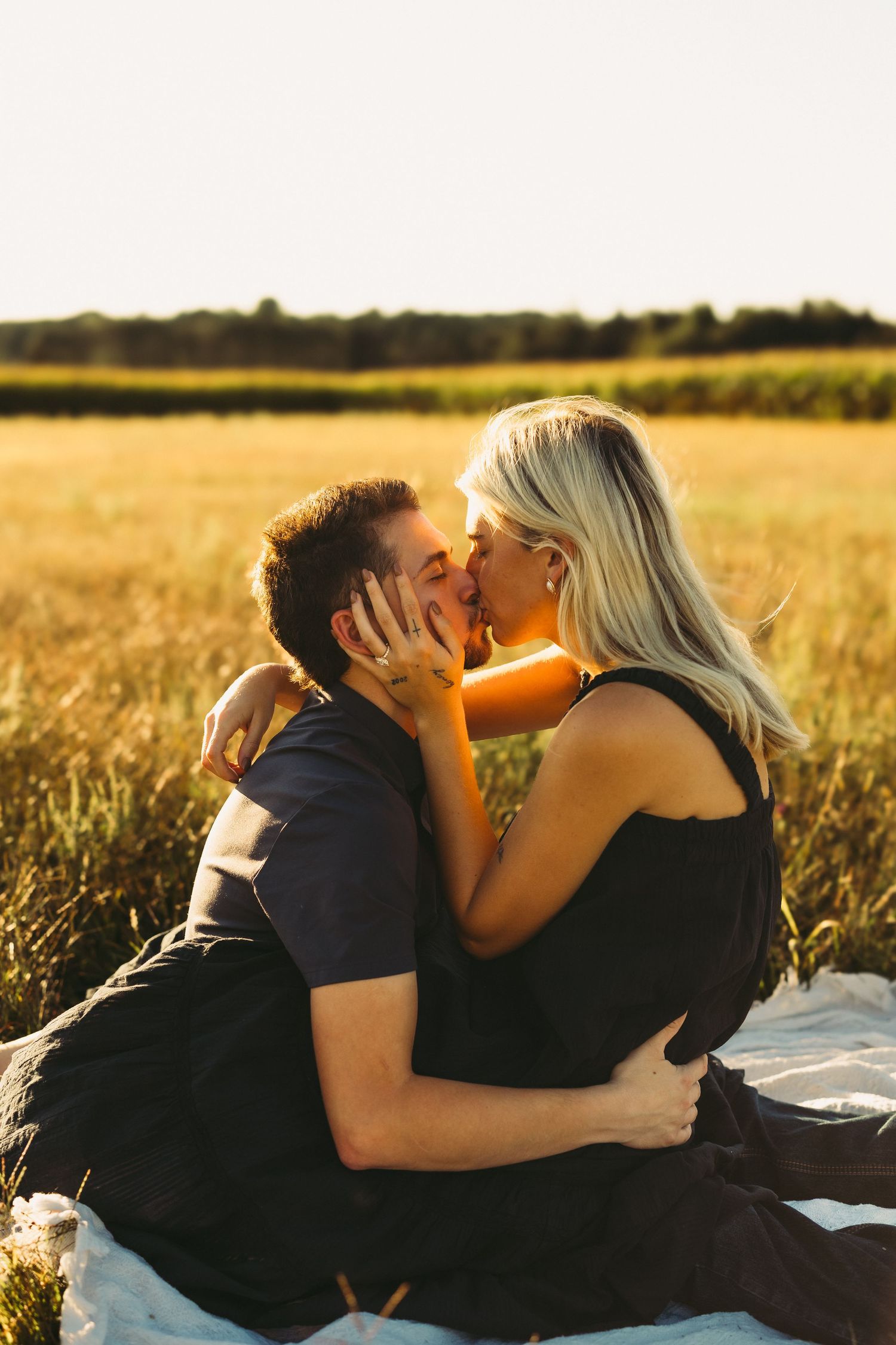 Couple sharing a romantic kiss on a blanket in a sunlit golden field, the woman in a black dress embracing the man.