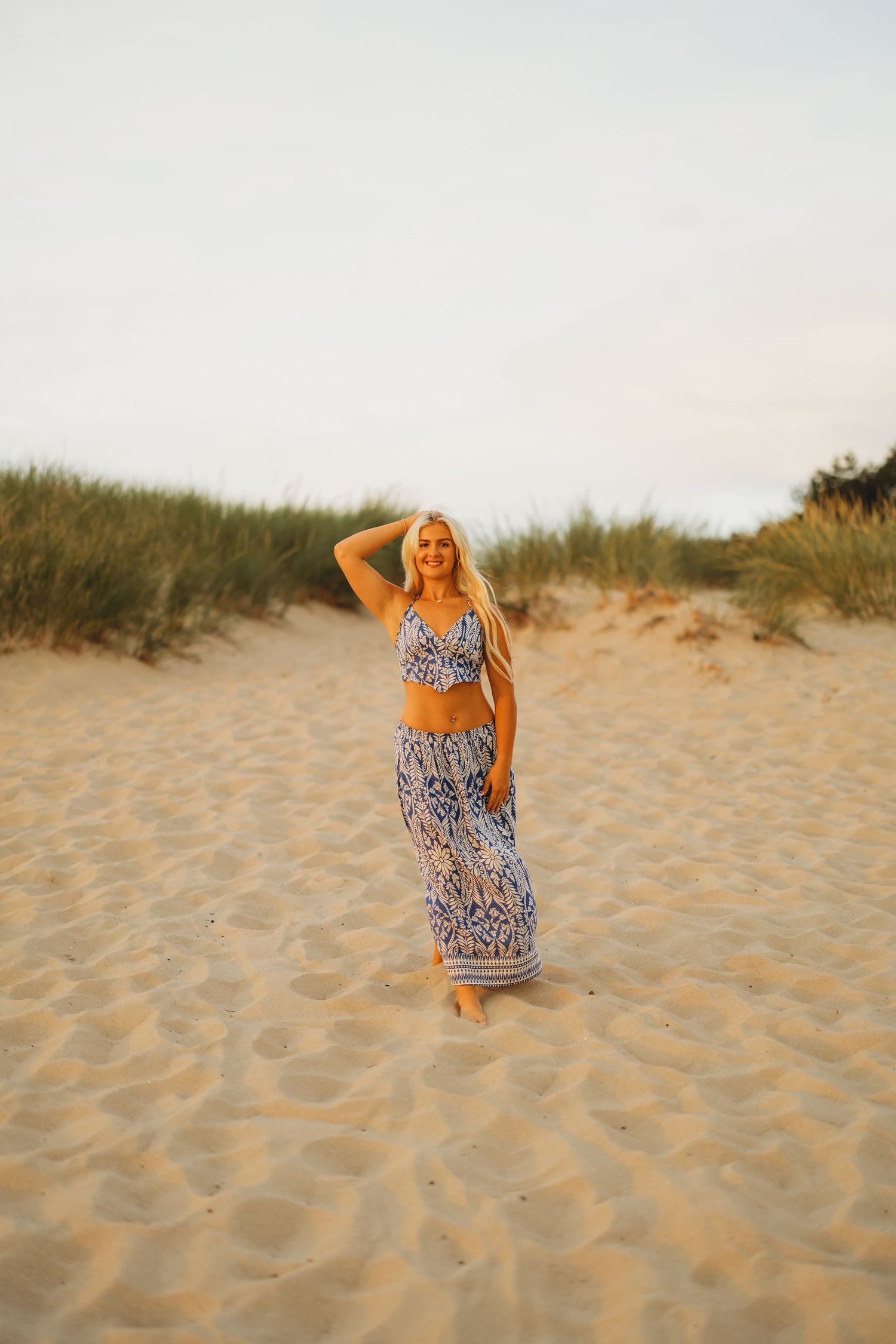 Blonde woman in a blue patterned two-piece outfit stands on a sandy beach dune with one arm raised above her head.