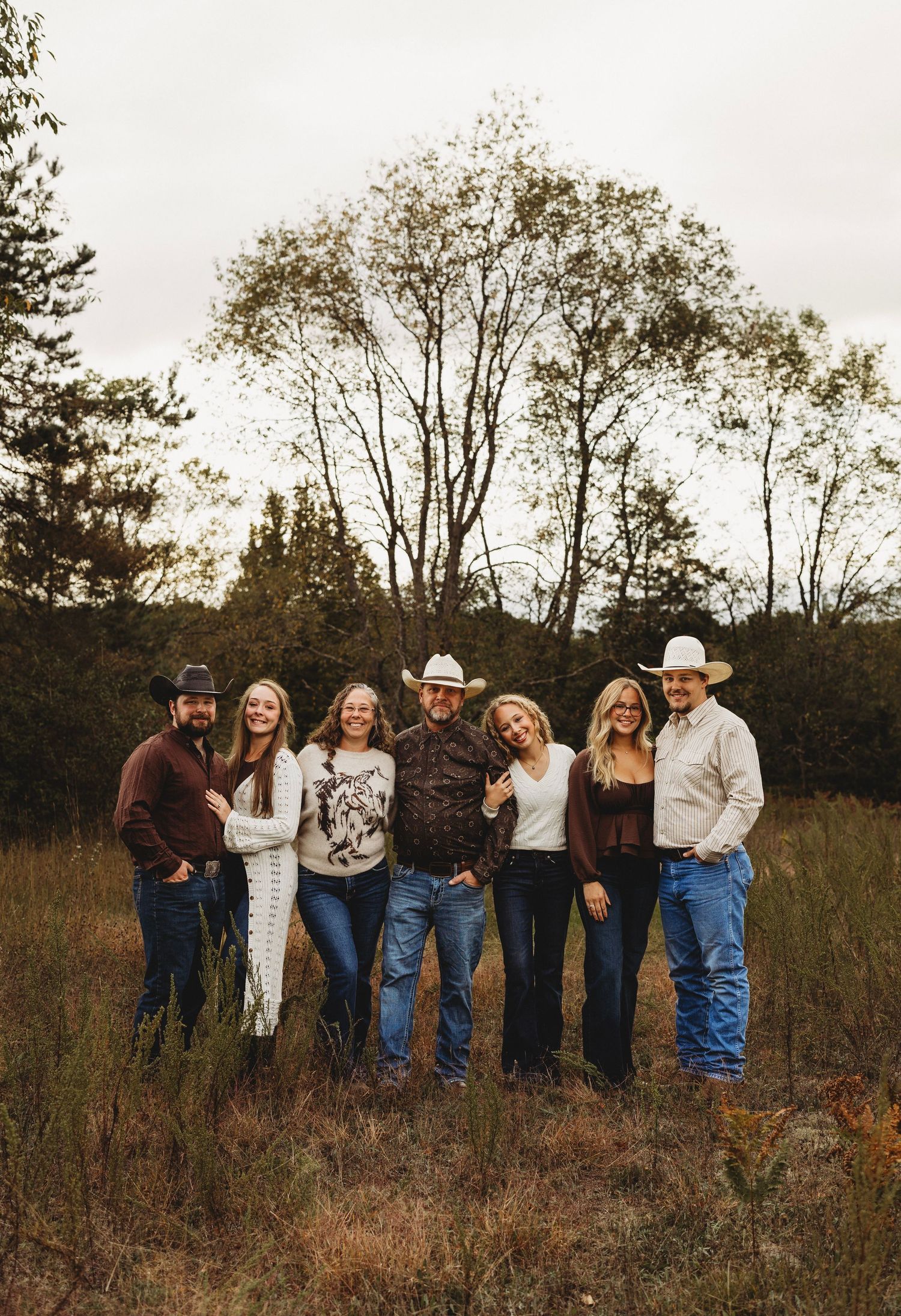 Six family members in cowboy hats and western clothing pose together in a golden autumn field with bare trees and cloudy skies.