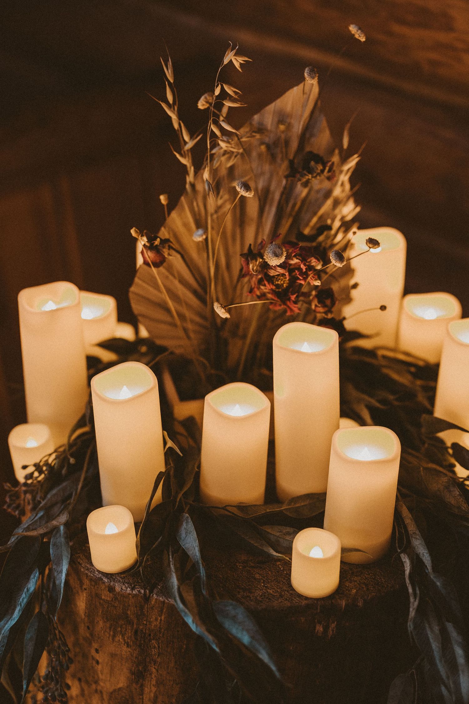 Multiple glowing LED candles arranged on a dark surface with dried palm leaves create a moody atmospheric setting.