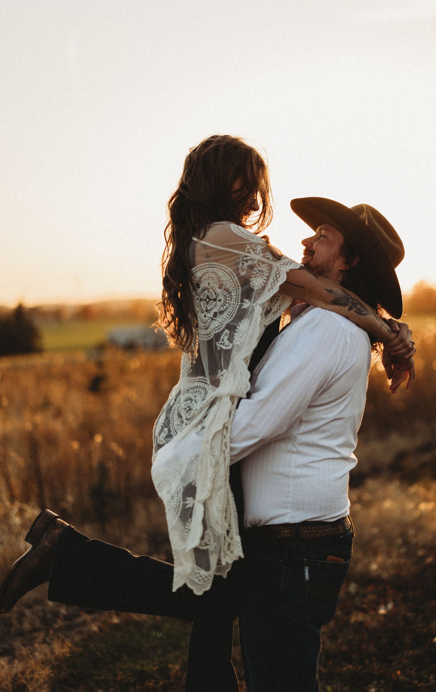 A romantic couple embraces in a golden field at sunset, with the bride wearing a flowing white vintage lace dress.