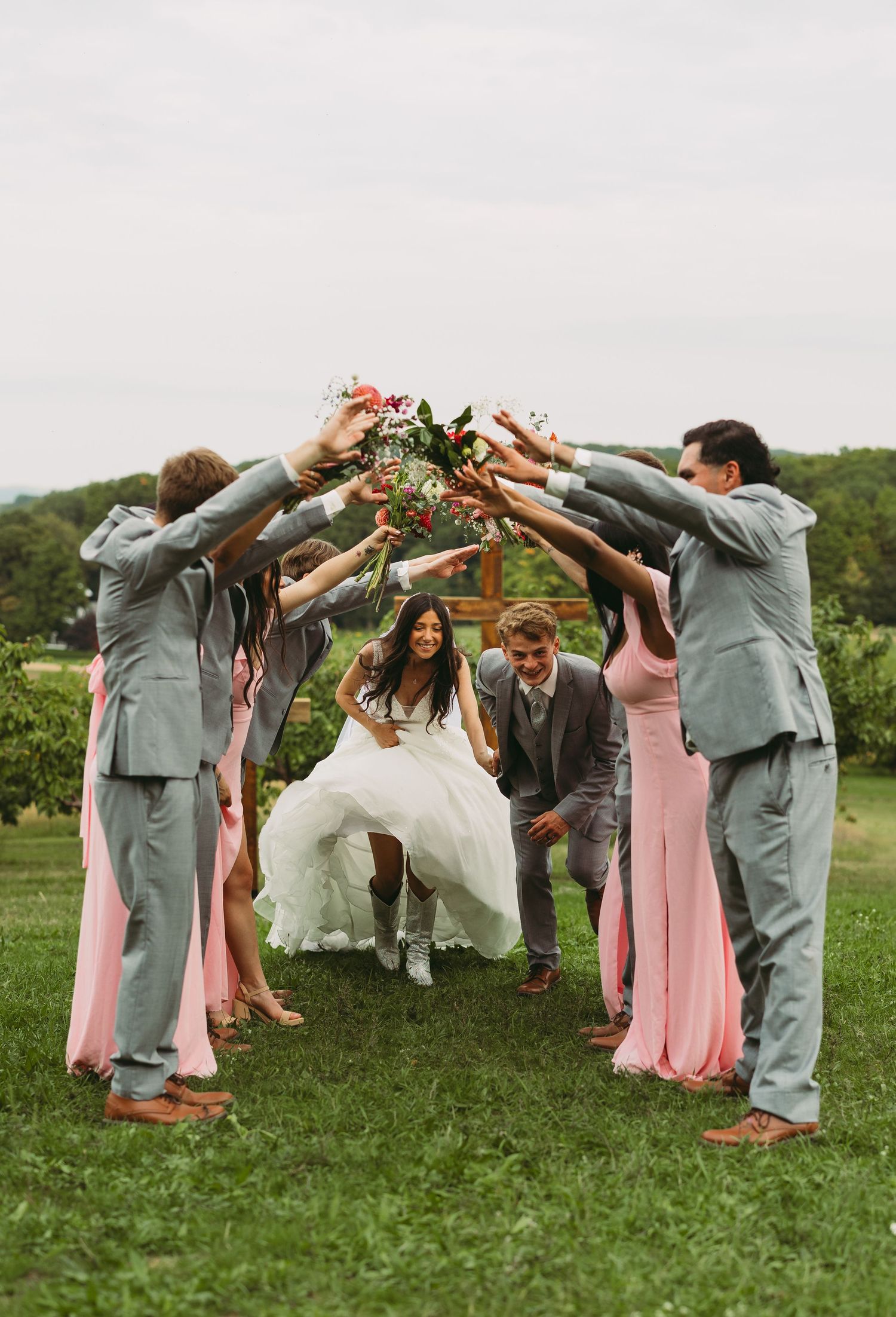 Wedding party forms a tunnel with raised arms for the newlyweds to walk through on a grassy field.