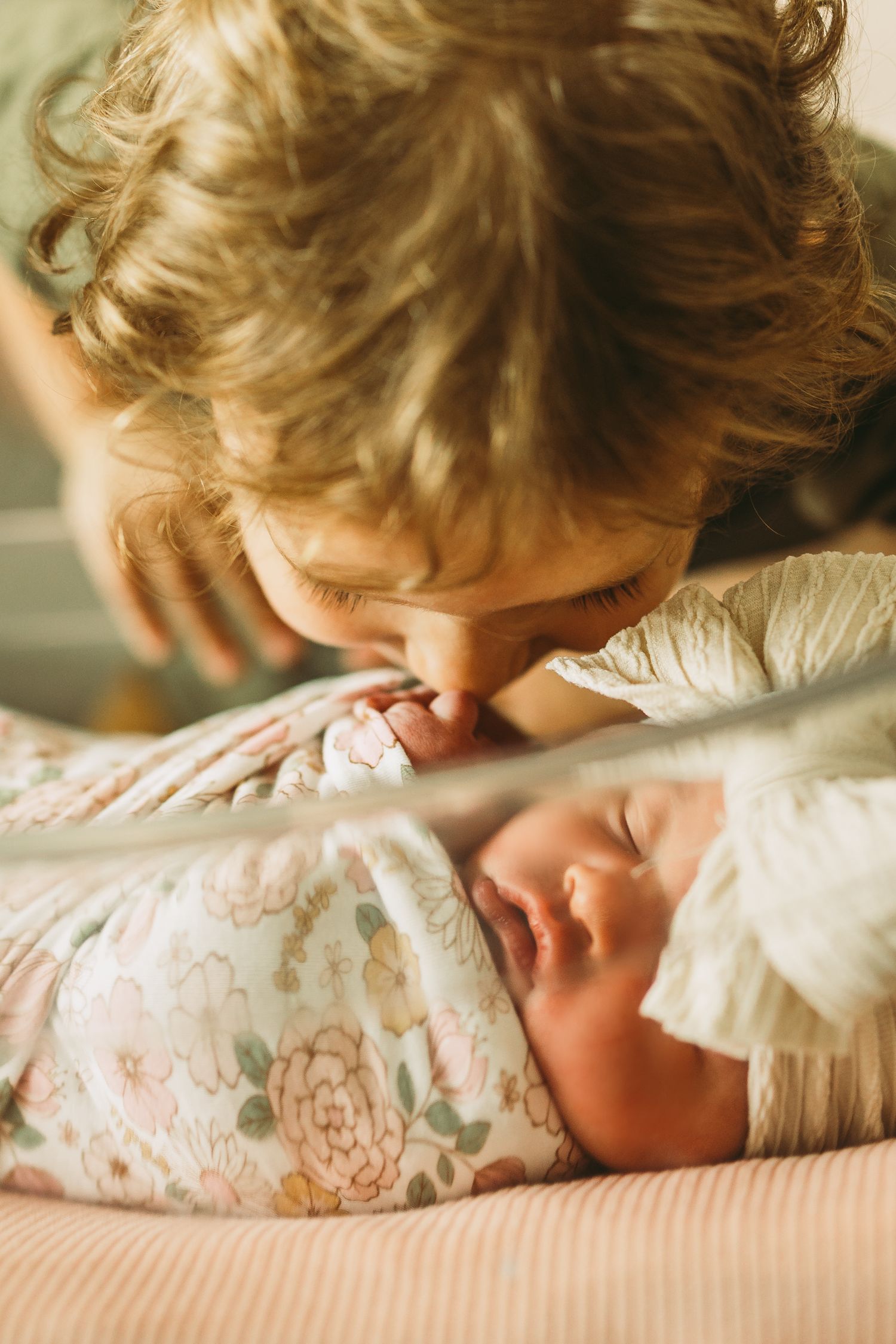 Intimate moment of sibling gently kissing newborn baby wrapped in floral blanket.