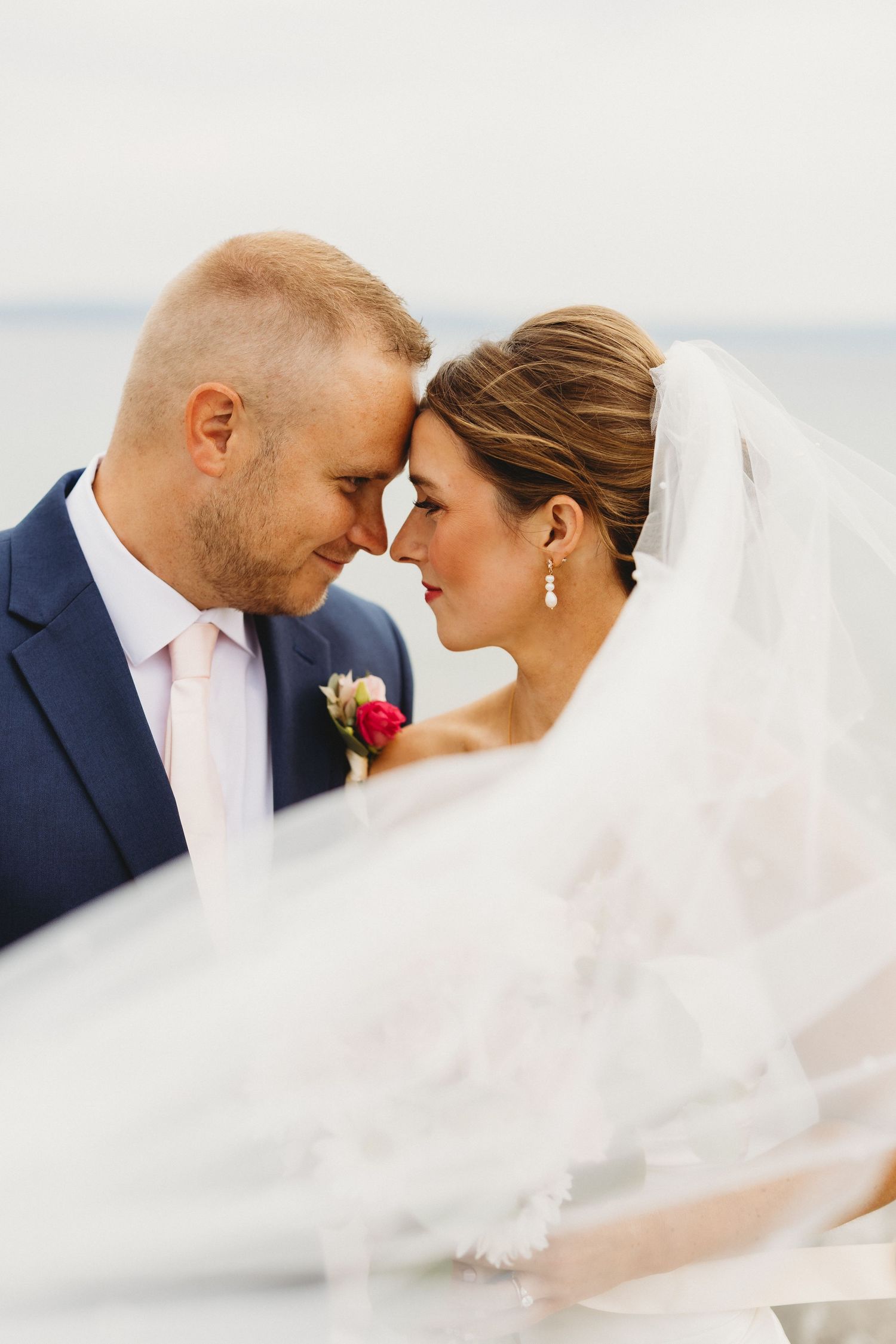 Bride and groom touching foreheads romantically near the ocean, with flowing white veil in foreground.