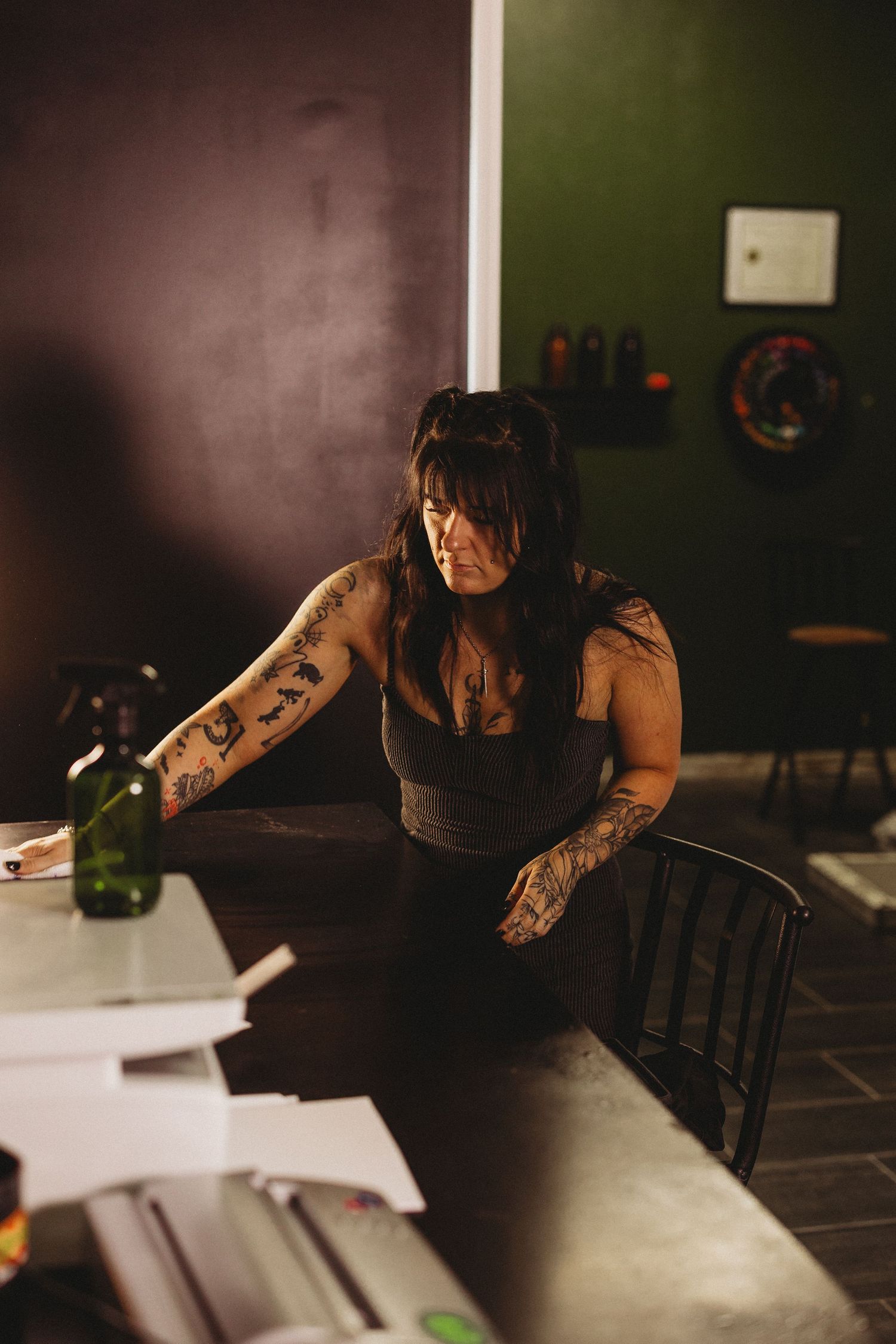 Tattooed woman in black tank top leaning over a counter in a dimly lit tattoo studio.