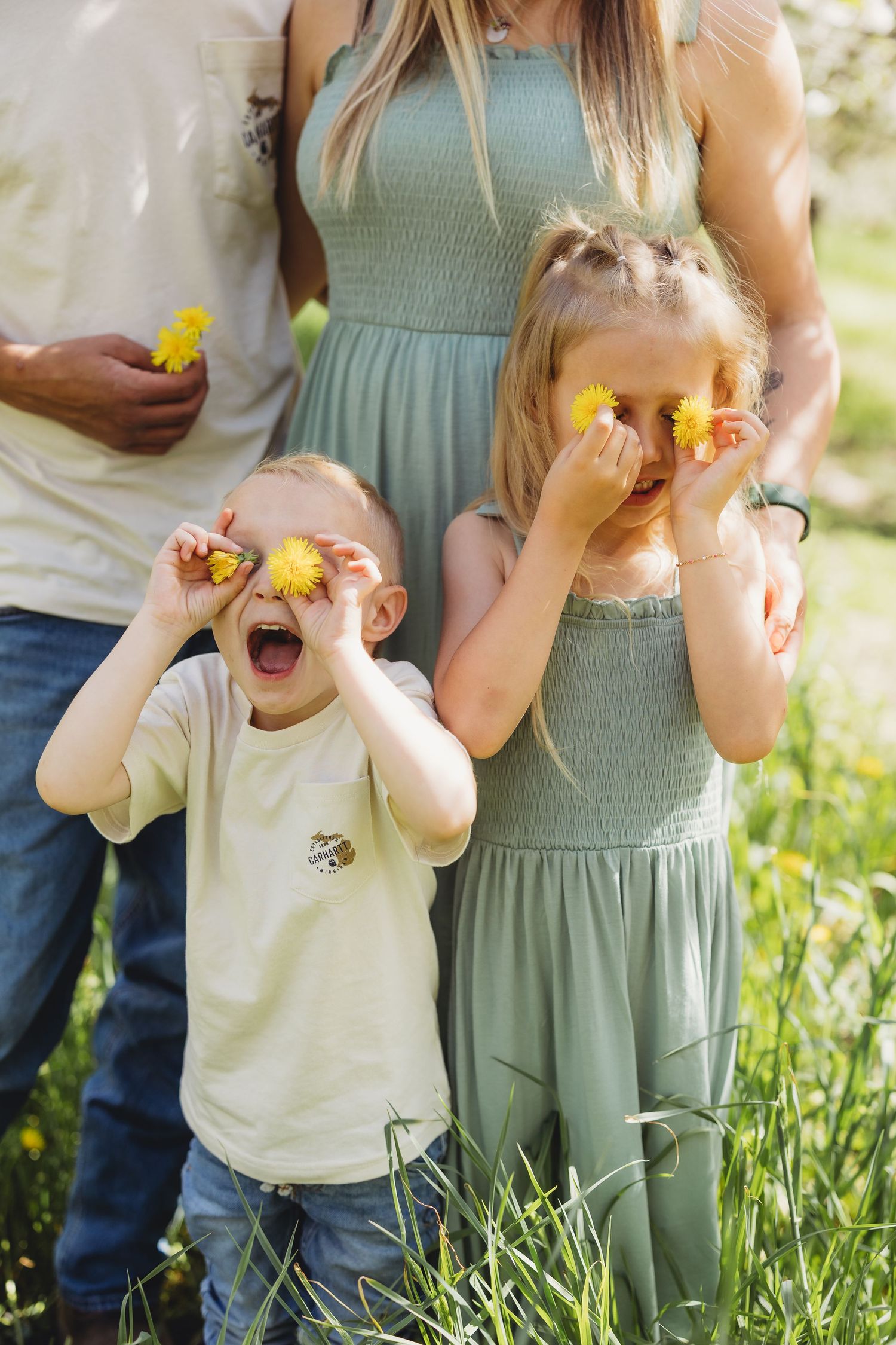 Two children playfully hold yellow dandelions over their eyes outdoors, standing with their parents in a sunny green field.