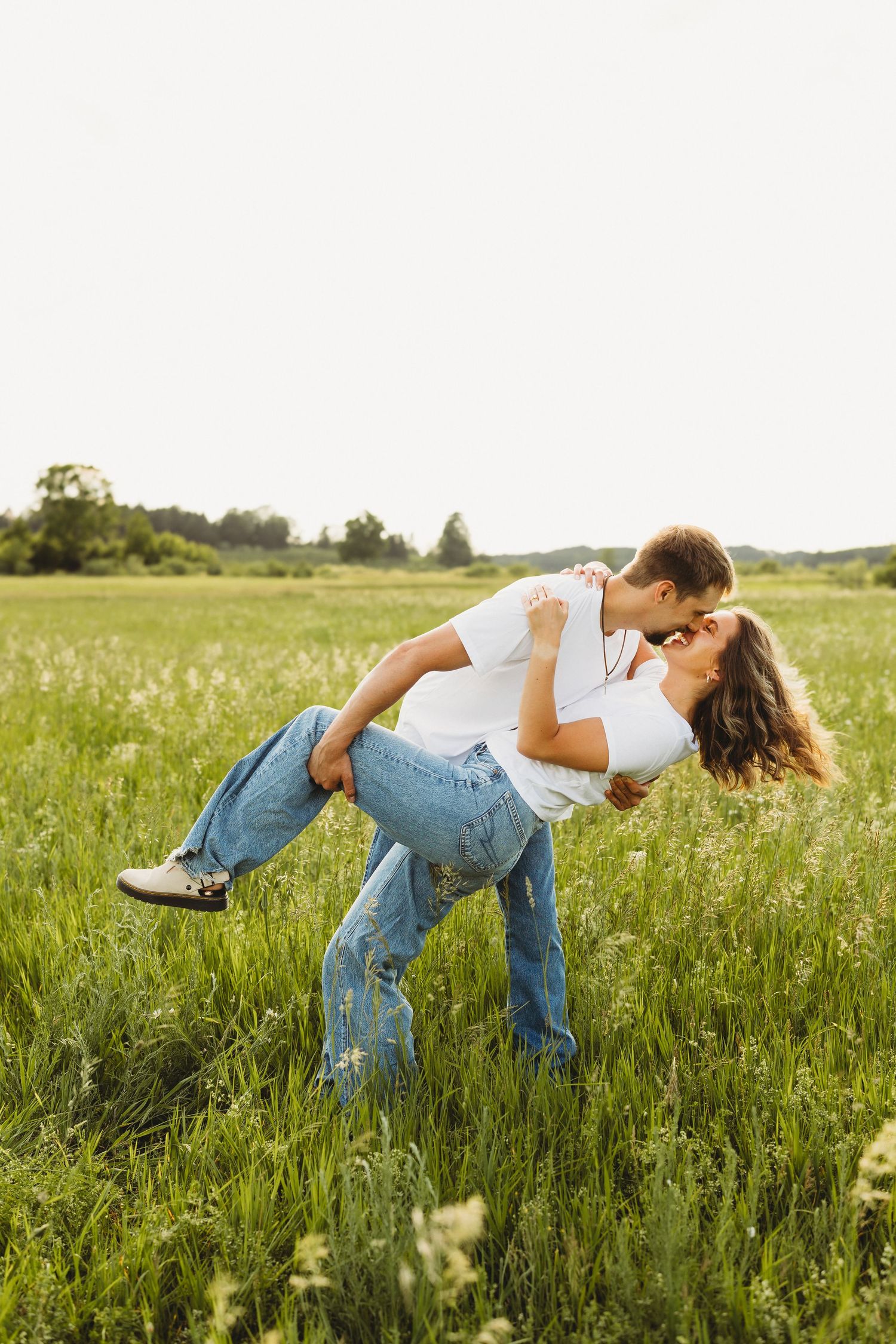 Couple sharing a romantic dip kiss in a lush green field, wearing white shirts and jeans on a sunny day.