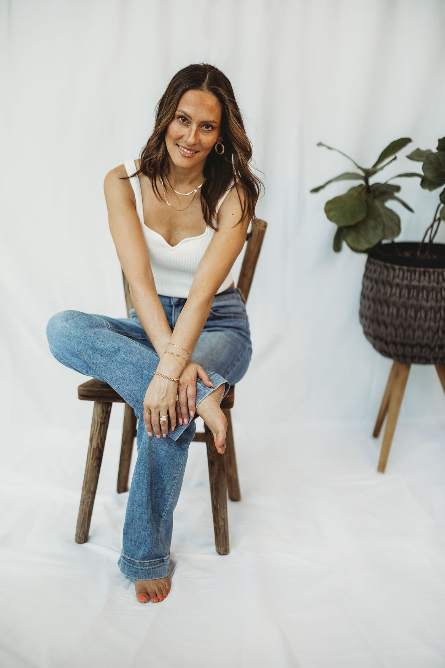 Woman in white top and jeans sitting on wooden chair, smiling, with a potted plant on white background.