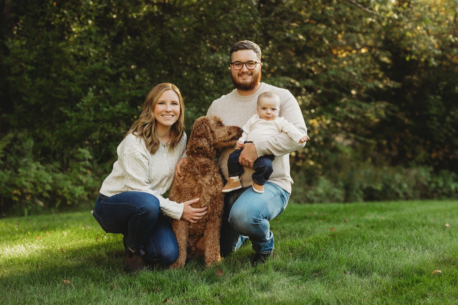 Family portrait with couple, baby, and golden doodle dog posing on green grass with lush trees in background.