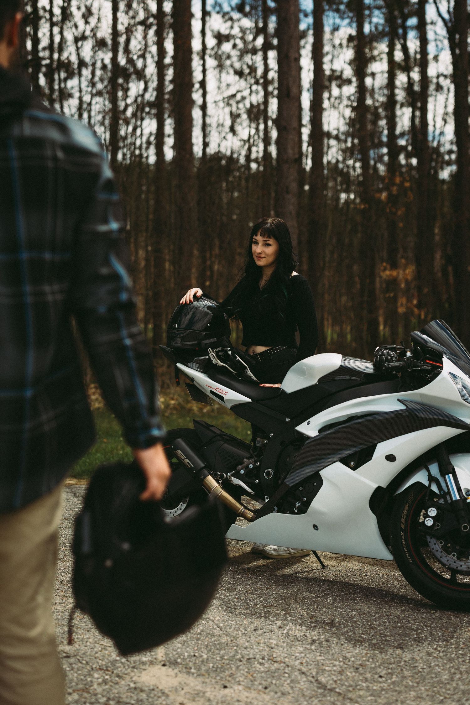 A woman in black sits on a white sport motorcycle as a man in plaid shirt approaches holding a helmet in a forest.