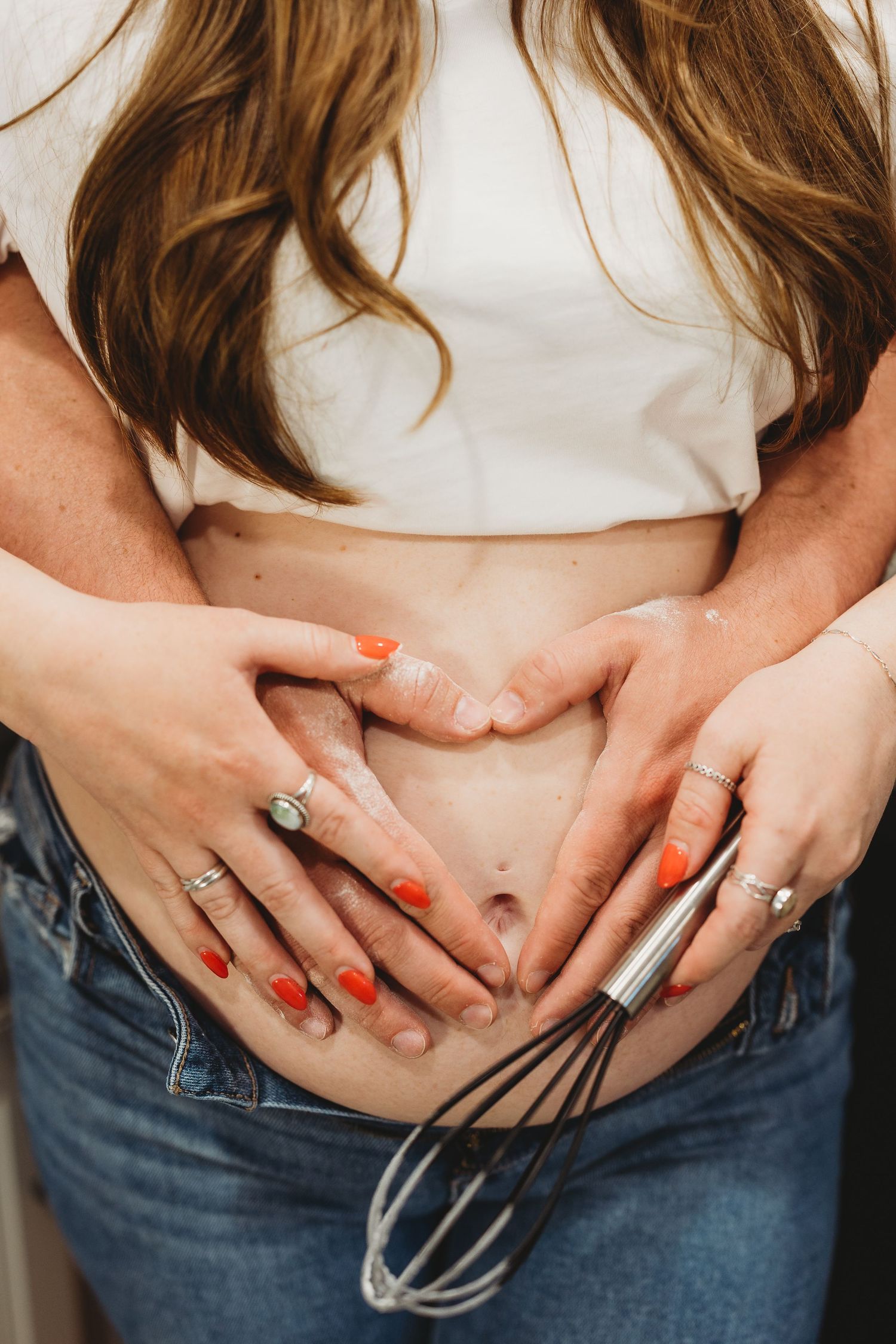 Pregnant woman in white crop top and jeans with couple forming heart shape on baby bump, holding a whisk.