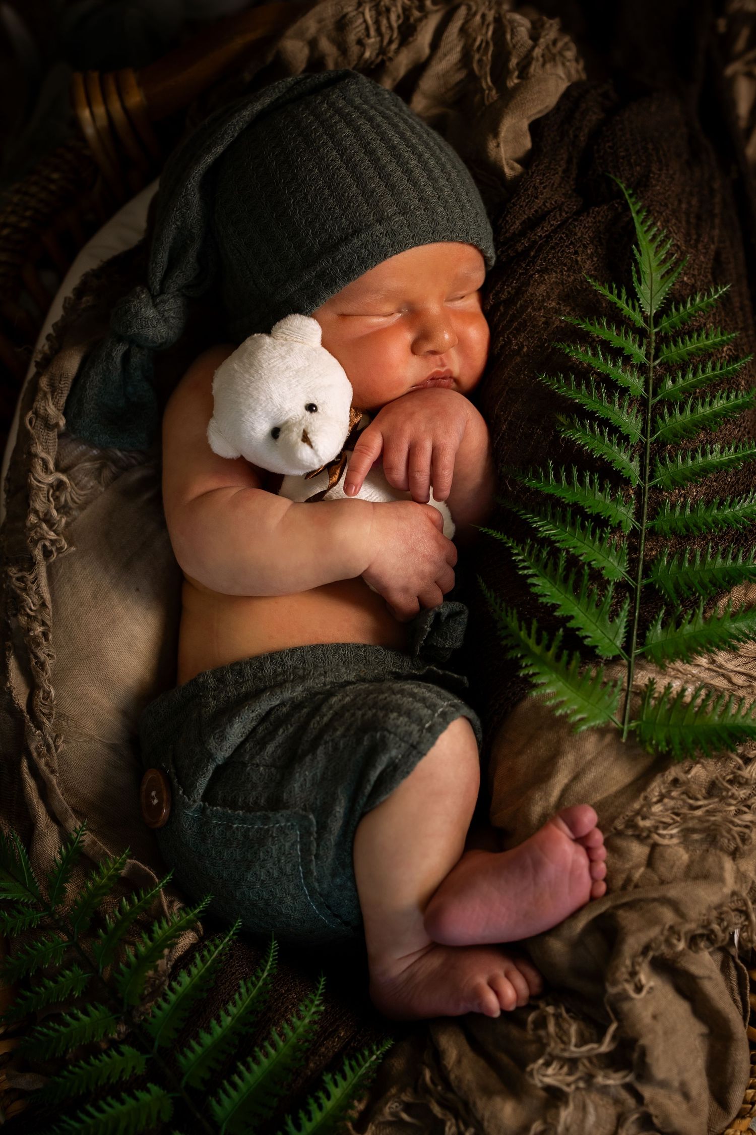 Sleeping newborn baby in green knit hat and shorts holding a white teddy bear, nestled in a basket with fern leaves.
