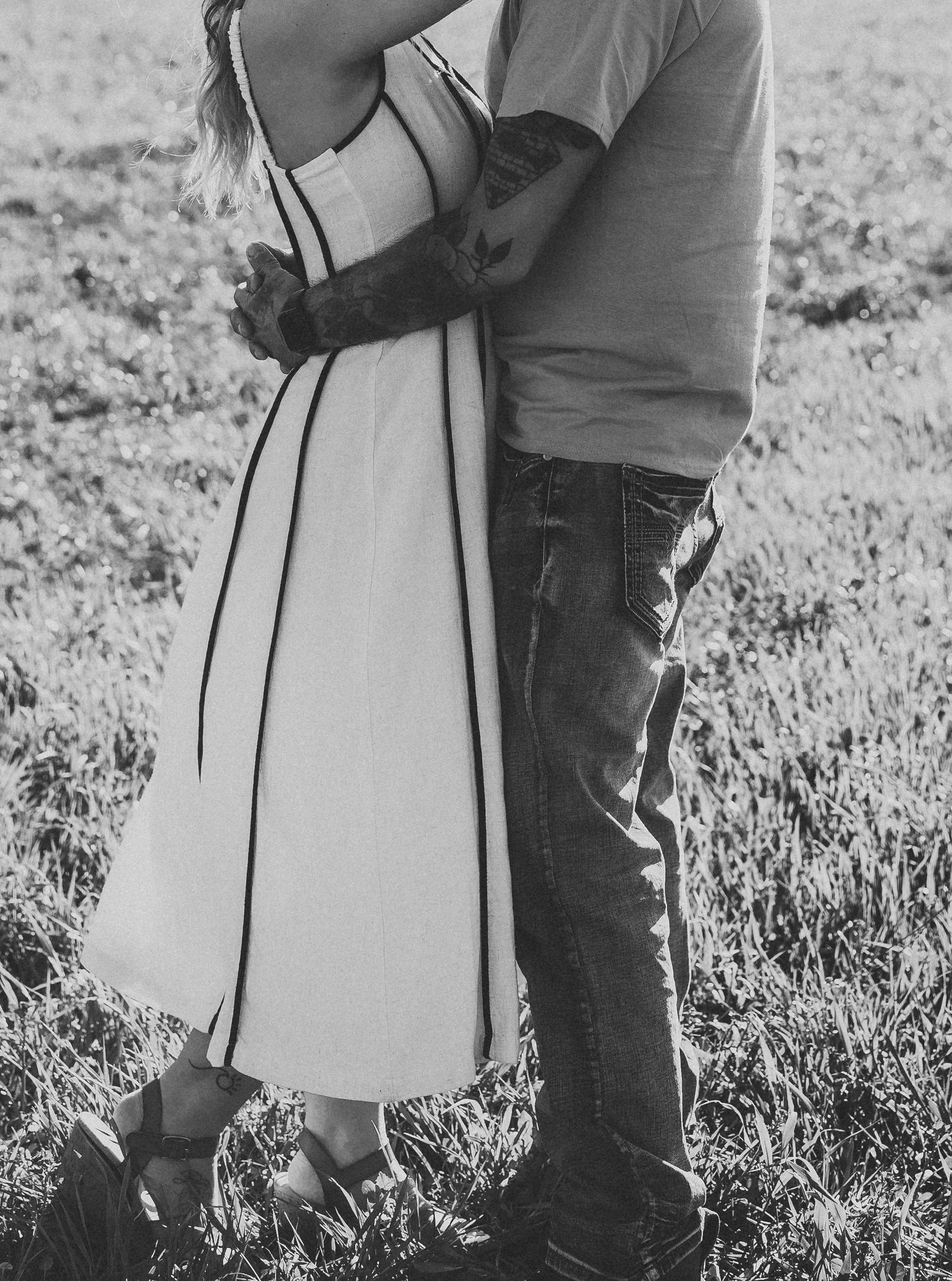 Black and white photo of a couple embracing in a field, woman in white dress with black trim, man in jeans.