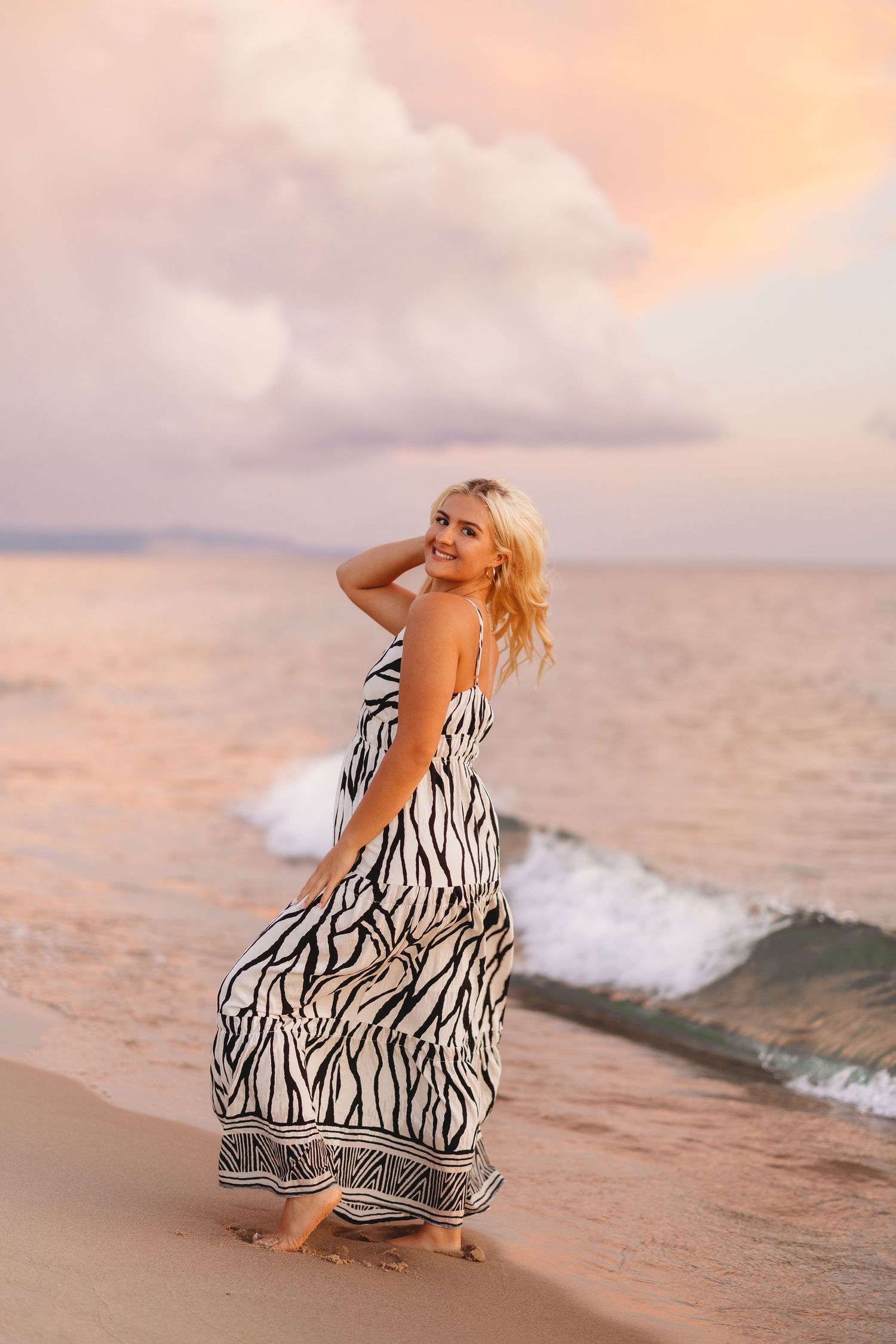 Smiling blonde woman in a zebra-print maxi dress standing barefoot at the water's edge as waves roll in under a pastel sunset sky.