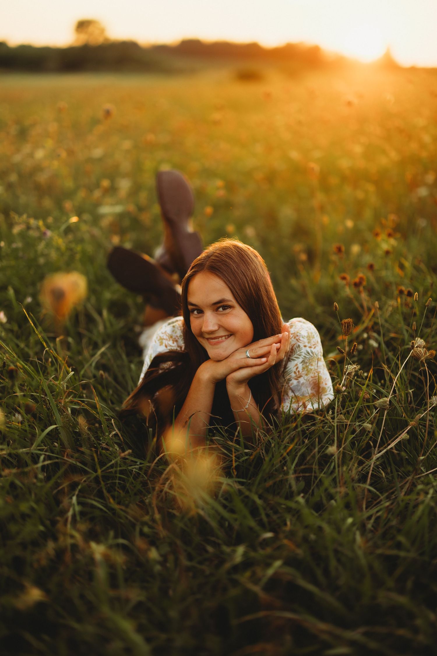 Smiling young woman with long brown hair lies in a wildflower field at golden hour, resting her chin on her hands with legs raised.