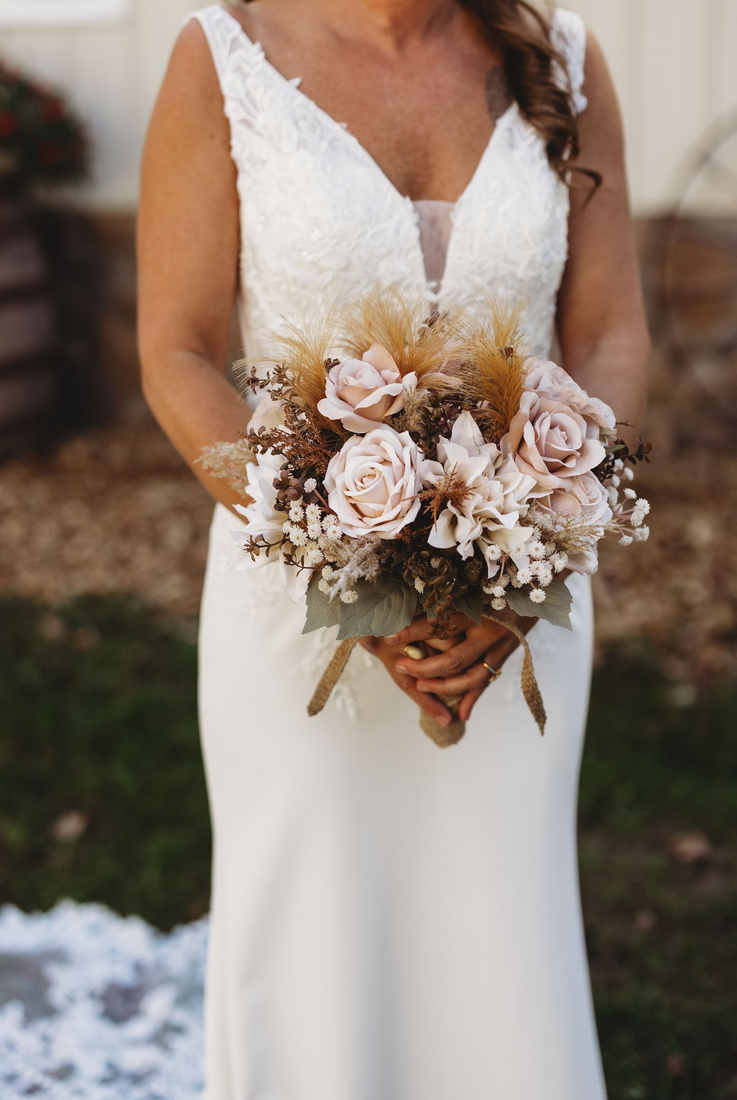 Bride in a white lace V-neck wedding gown holds a boho bouquet of blush roses, dried pampas grass, and baby's breath outdoors.