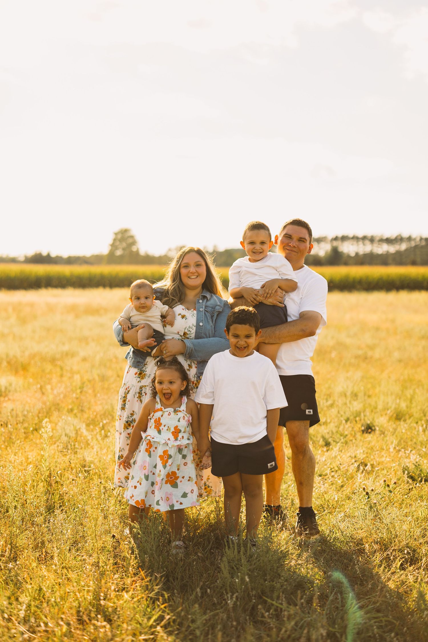 Smiling family of five with two parents and three young children posing together in a golden sunlit field at sunset.