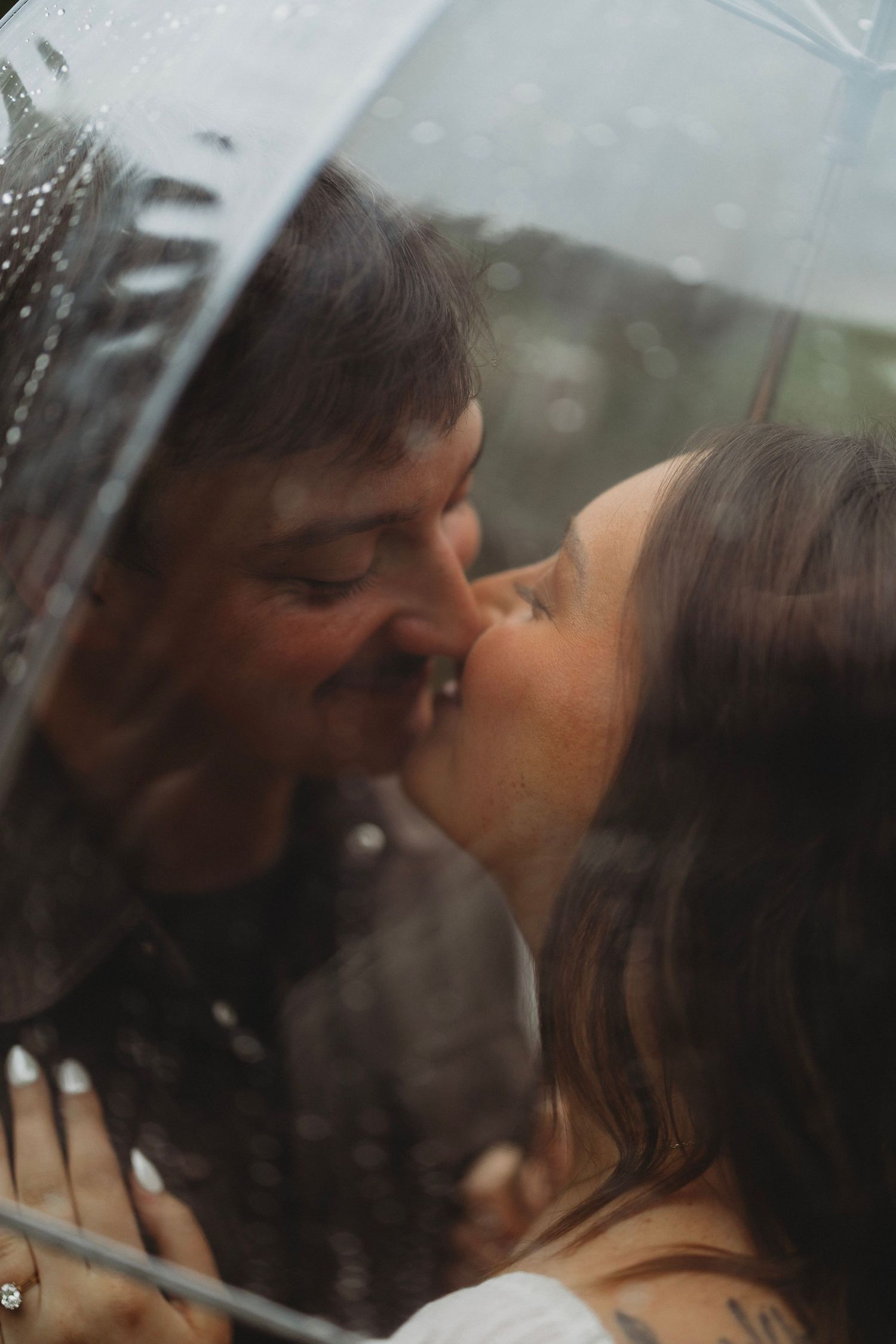 Couple sharing a romantic kiss under a clear umbrella dotted with raindrops, with blurred green trees in the background.