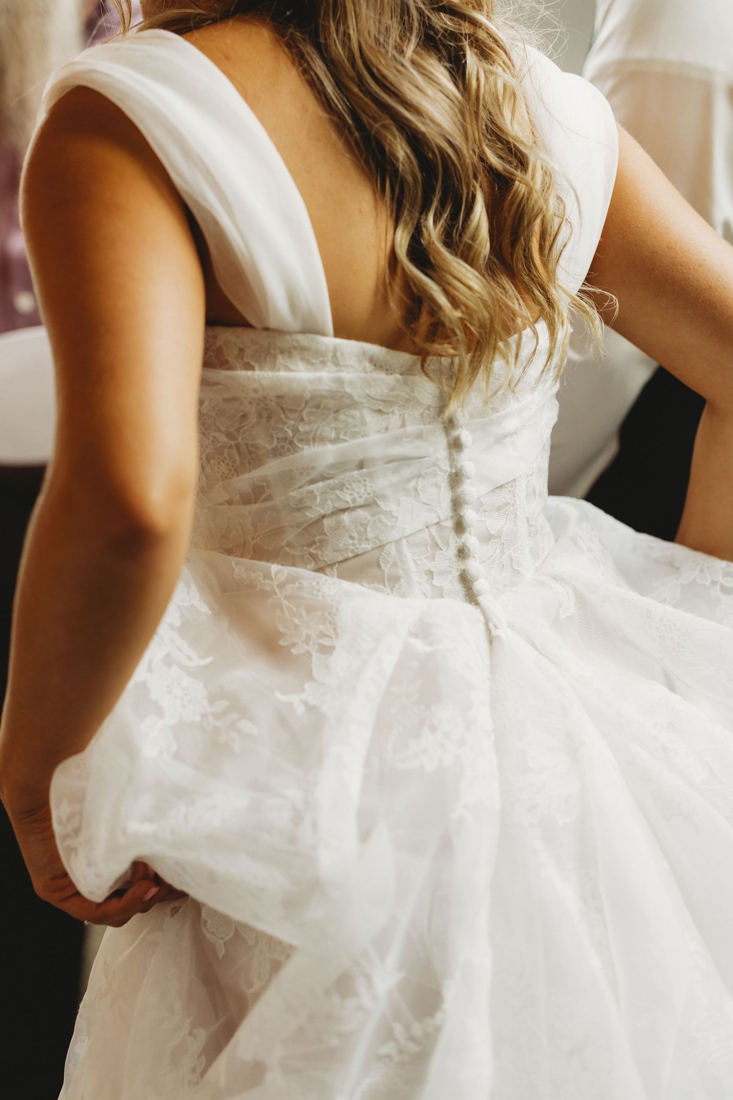 Back view of a bride in a white lace wedding gown with wide straps, covered buttons and long wavy blonde hair, holding her skirt.
