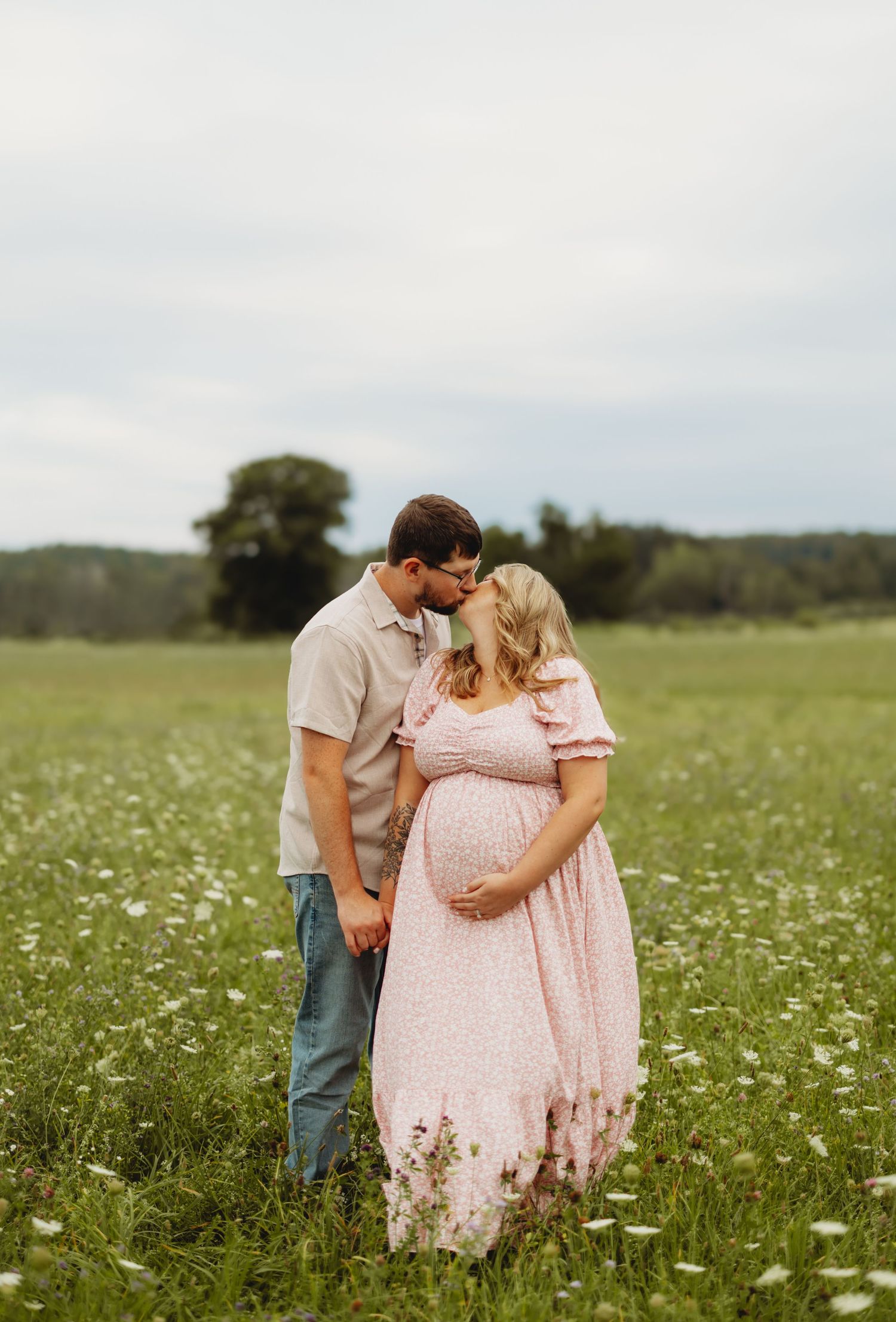 Couple sharing a kiss in wildflower meadow during maternity photo shoot, woman in pink floral dress.