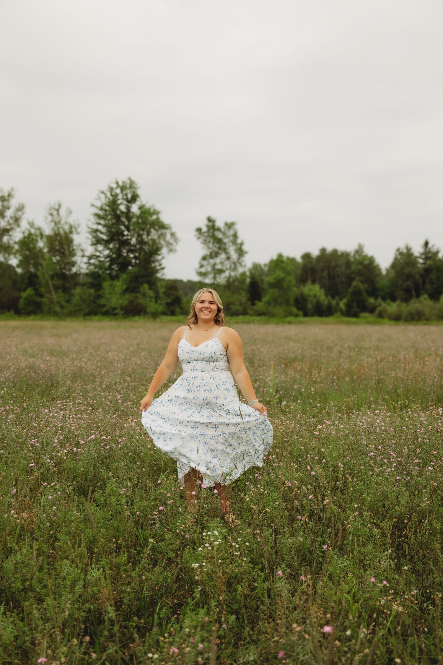 Woman in floral sundress twirling in a wildflower meadow with green trees in the background on a cloudy day.