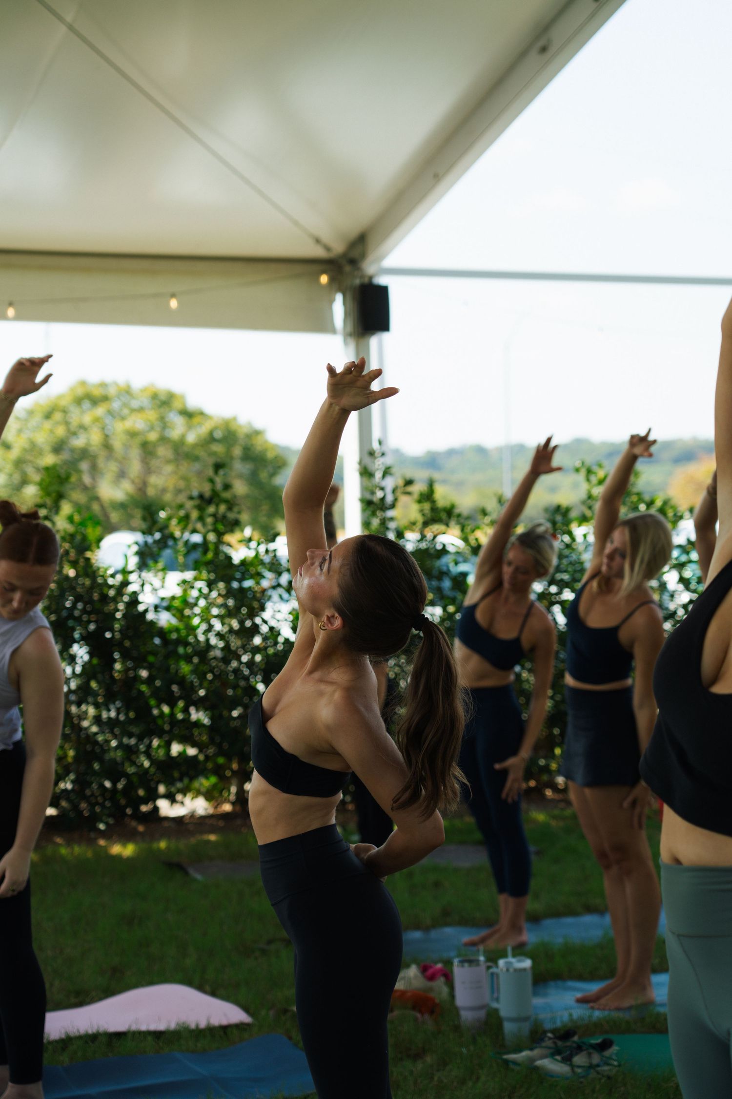 Women practicing outdoor yoga under a tent canopy, stretching arms upward in a group fitness class on colorful mats.