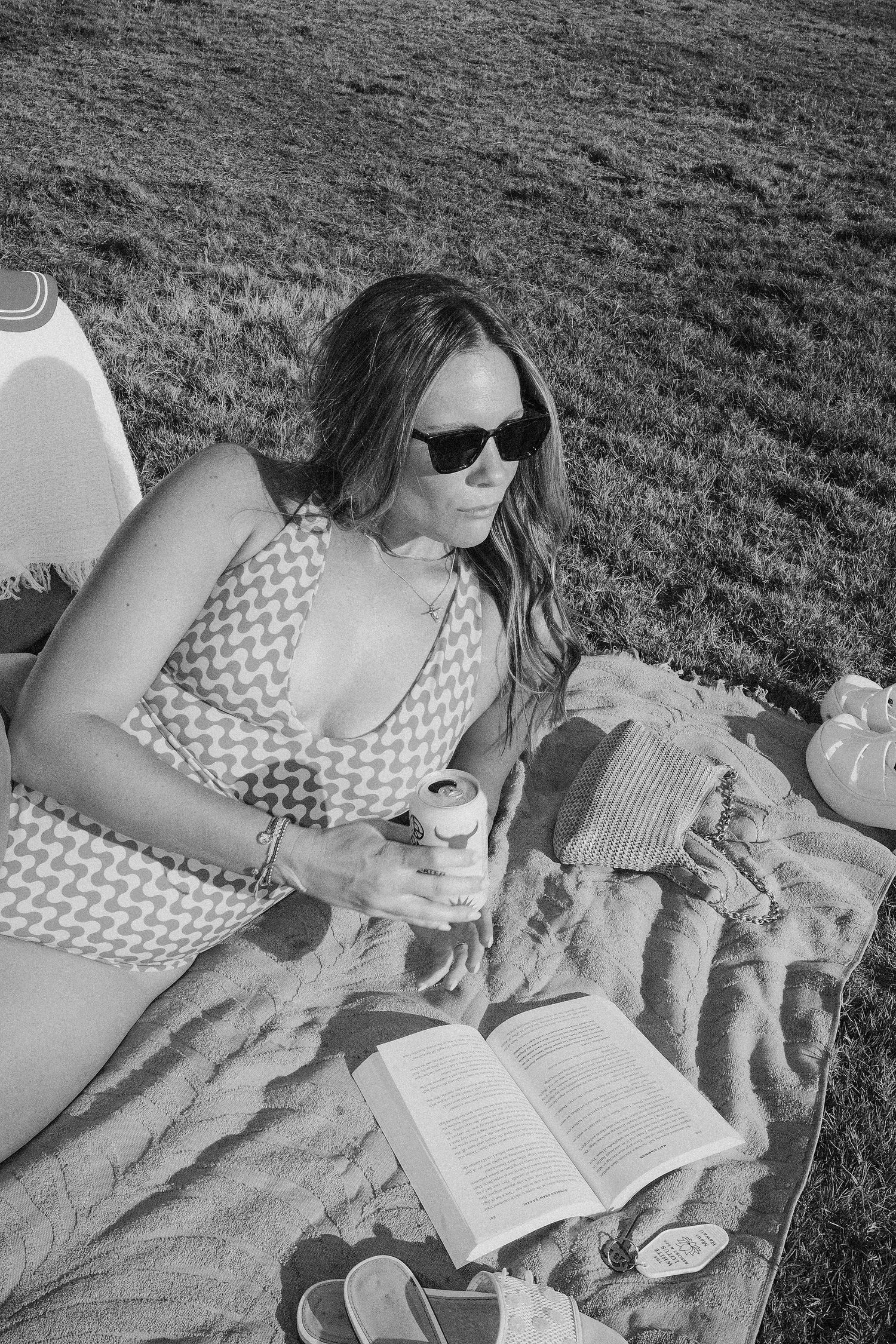 Woman in swimsuit reading a book on a blanket outdoors, holding a can, wearing sunglasses on a sunny day.