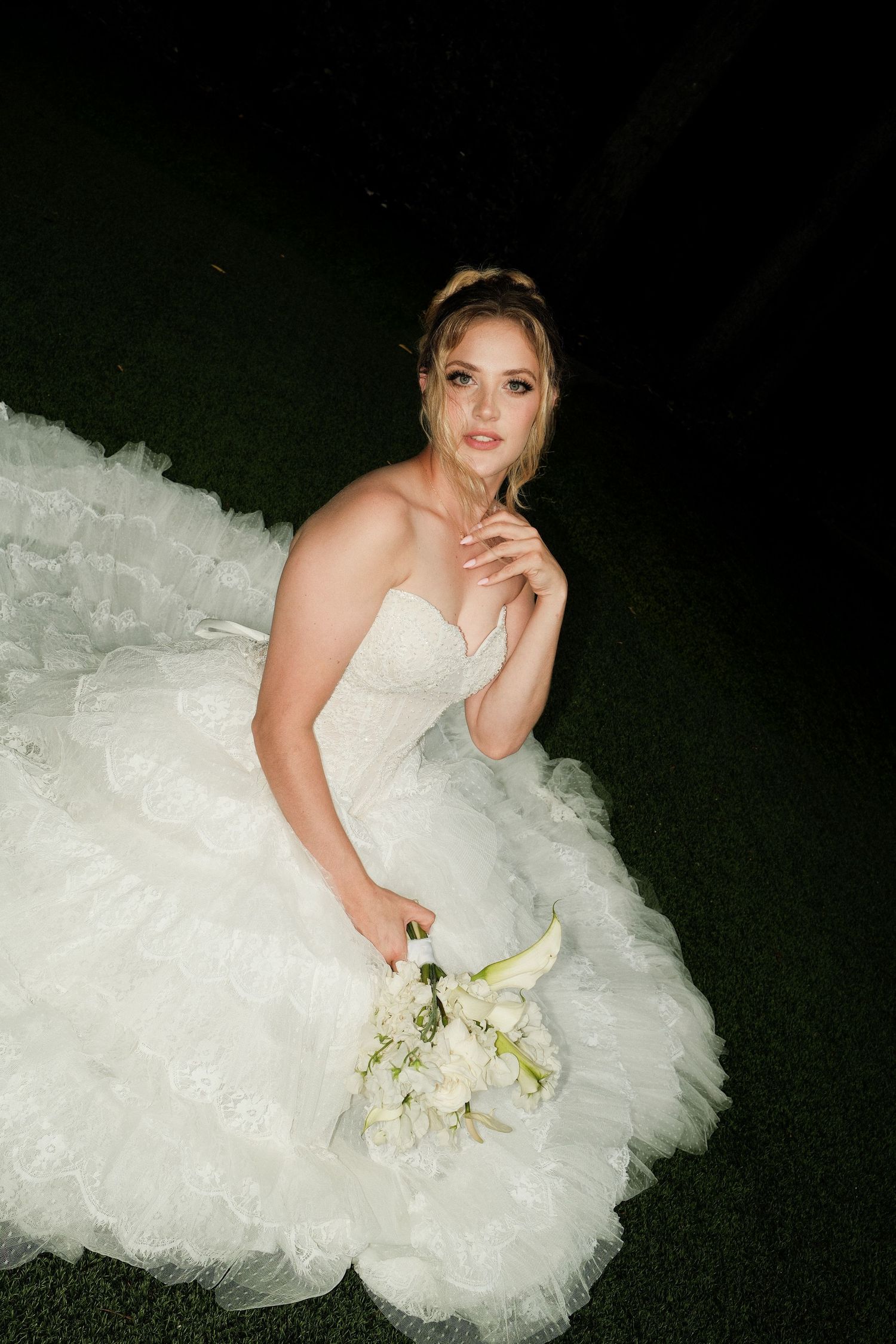 Bride in a strapless white ruffled ball gown sits on grass at night, holding a white floral bouquet.