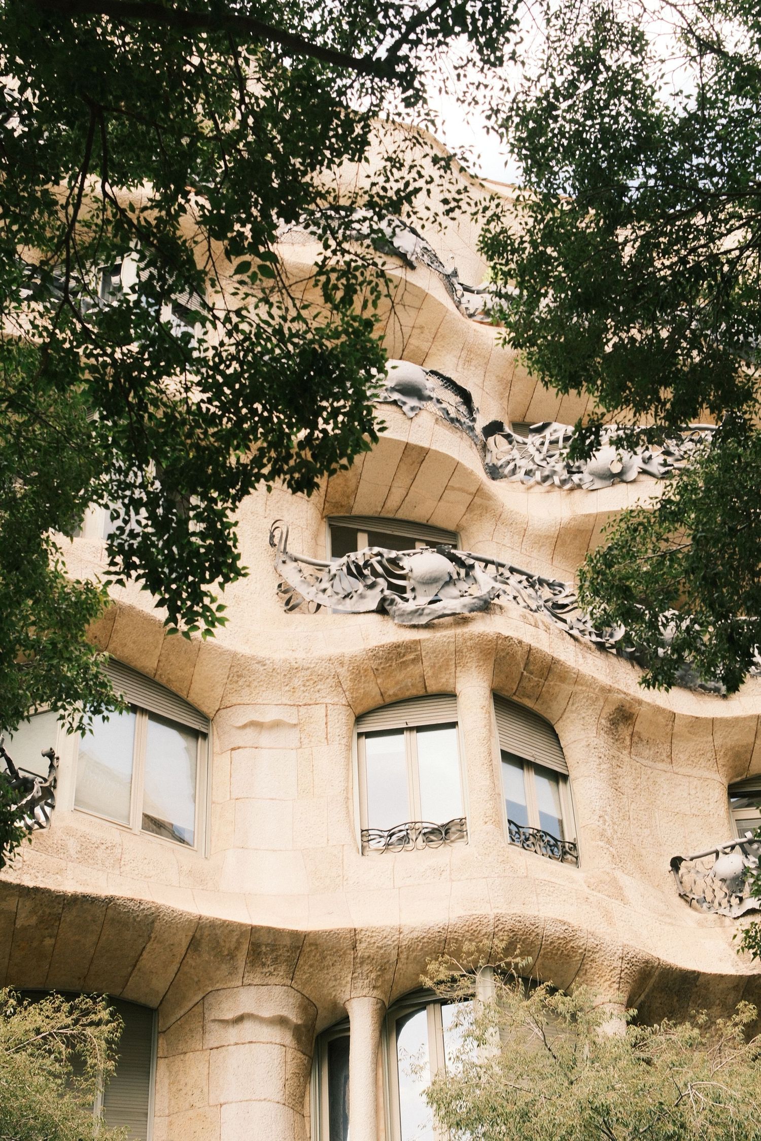 Curved facade of Casa Mila by Gaudi in Barcelona, featuring organic stone balconies and sculptural details framed by green trees.
