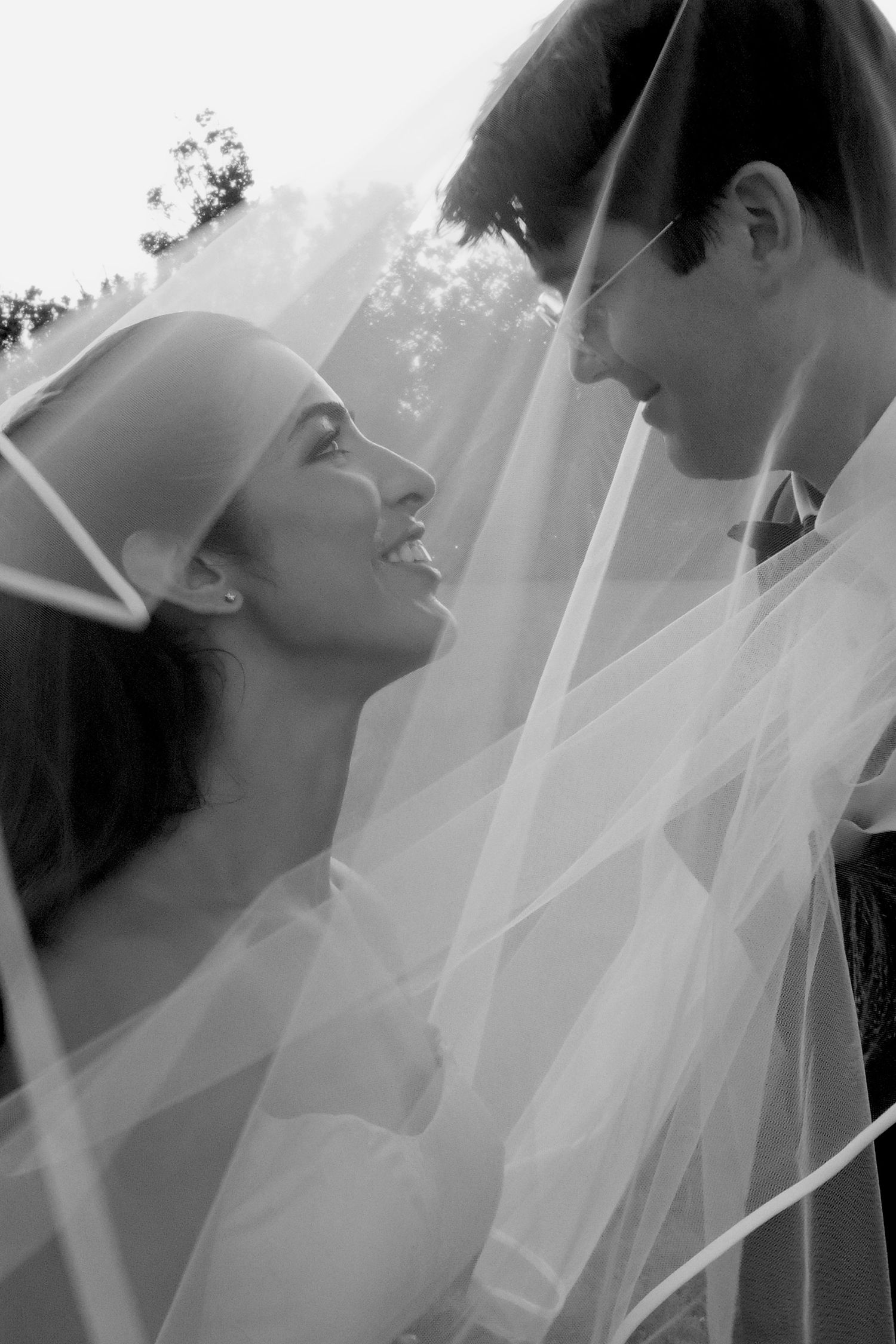 Black and white photo of a smiling bride and groom sharing a romantic moment under a flowing wedding veil outdoors.