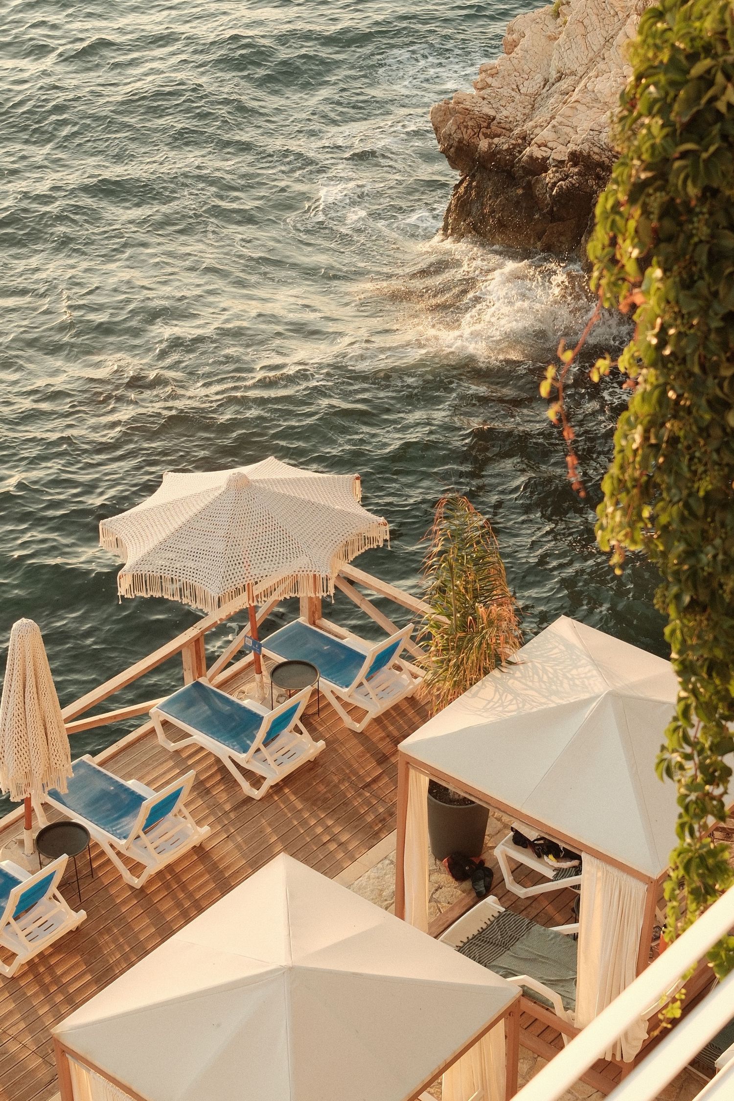 Aerial view of a luxury seaside terrace with blue lounge chairs, white umbrellas, and cabanas overlooking rocky ocean waters.