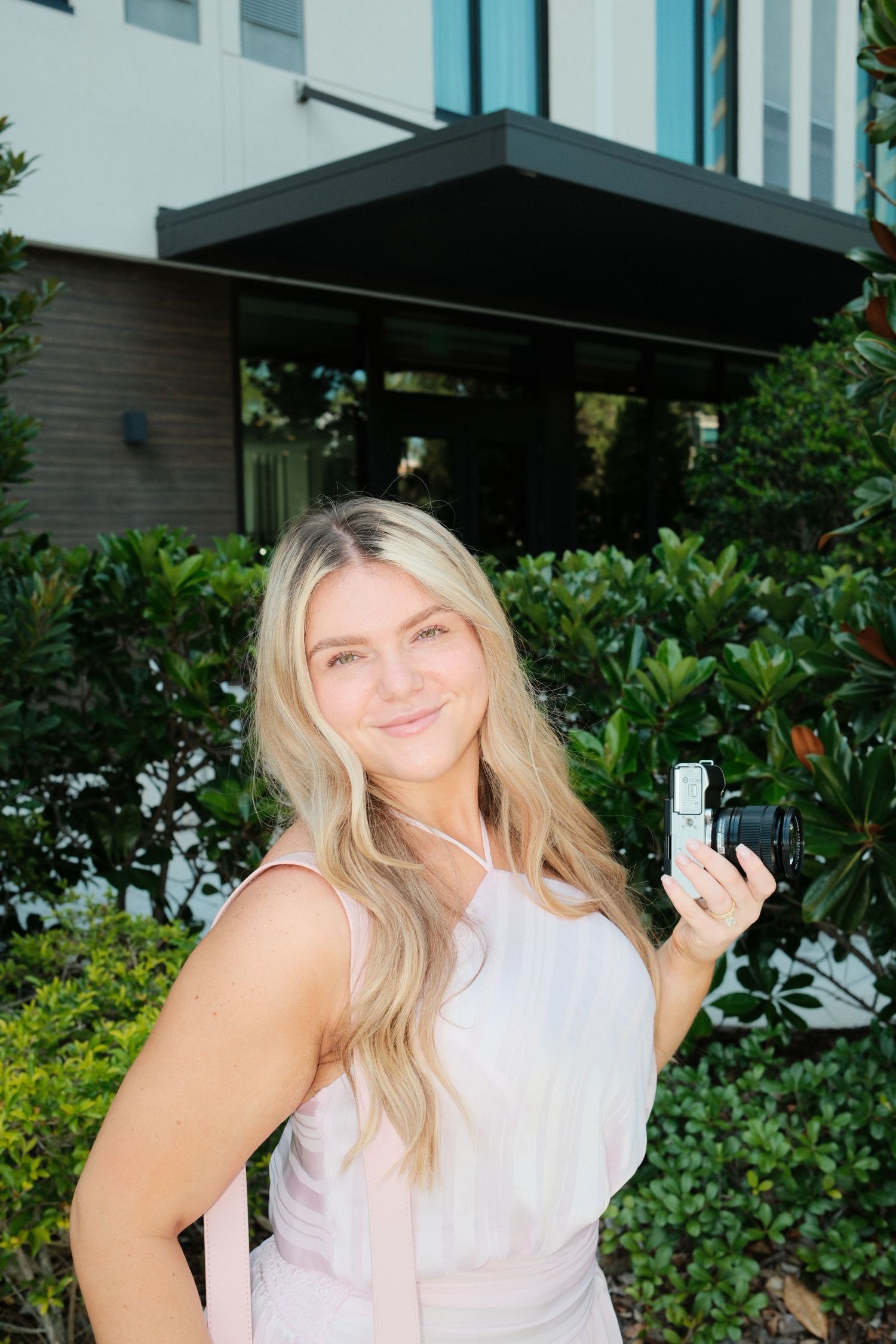 Smiling blonde woman in pink dress holding a camera outdoors near green shrubs and modern building entrance.