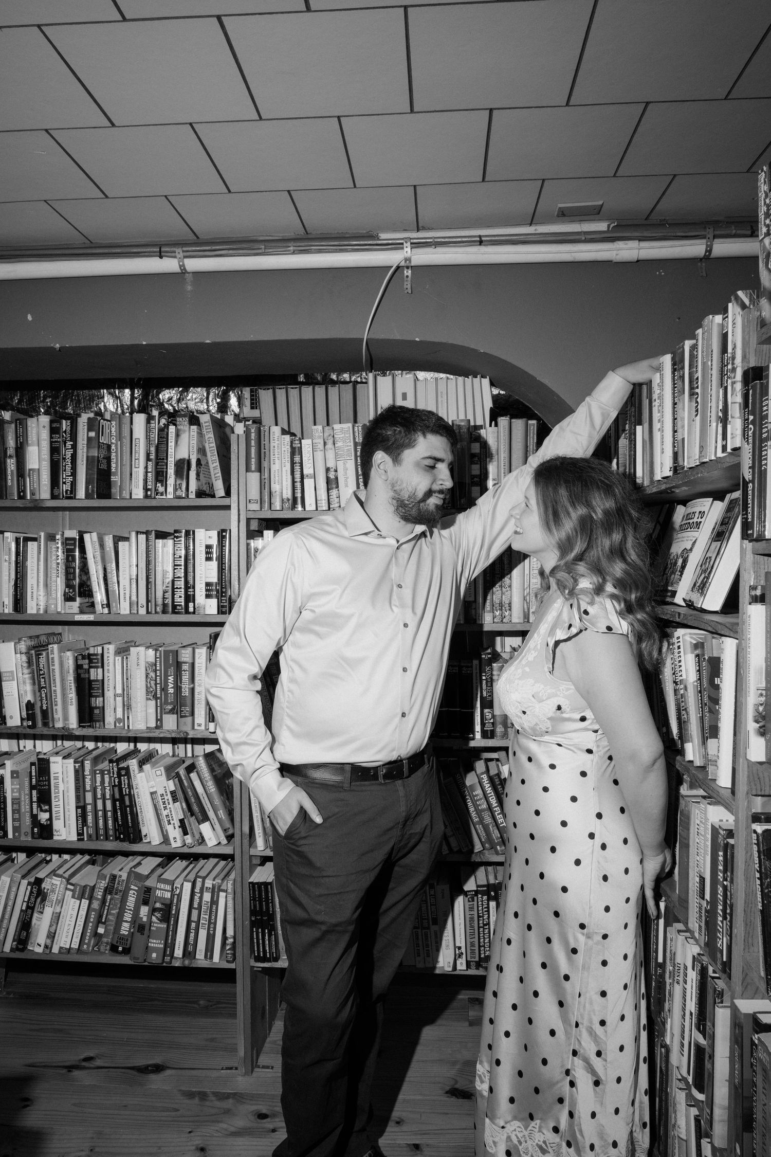 Black and white photo of a couple sharing a romantic moment between tall bookshelves in a cozy library or bookstore.
