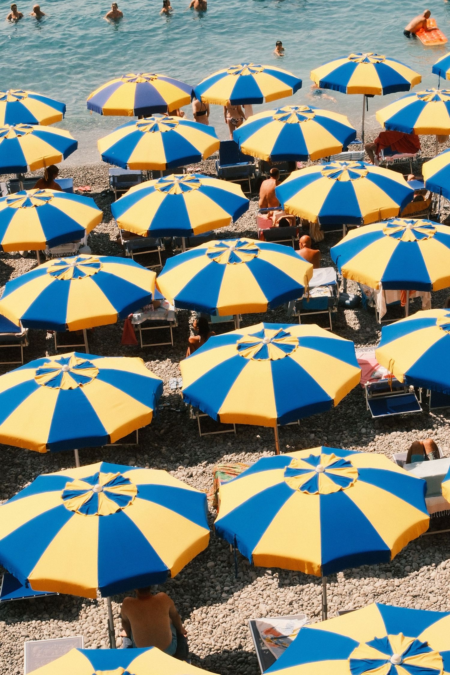 Aerial view of crowded beach with rows of blue and yellow striped umbrellas on pebble shore near blue ocean.