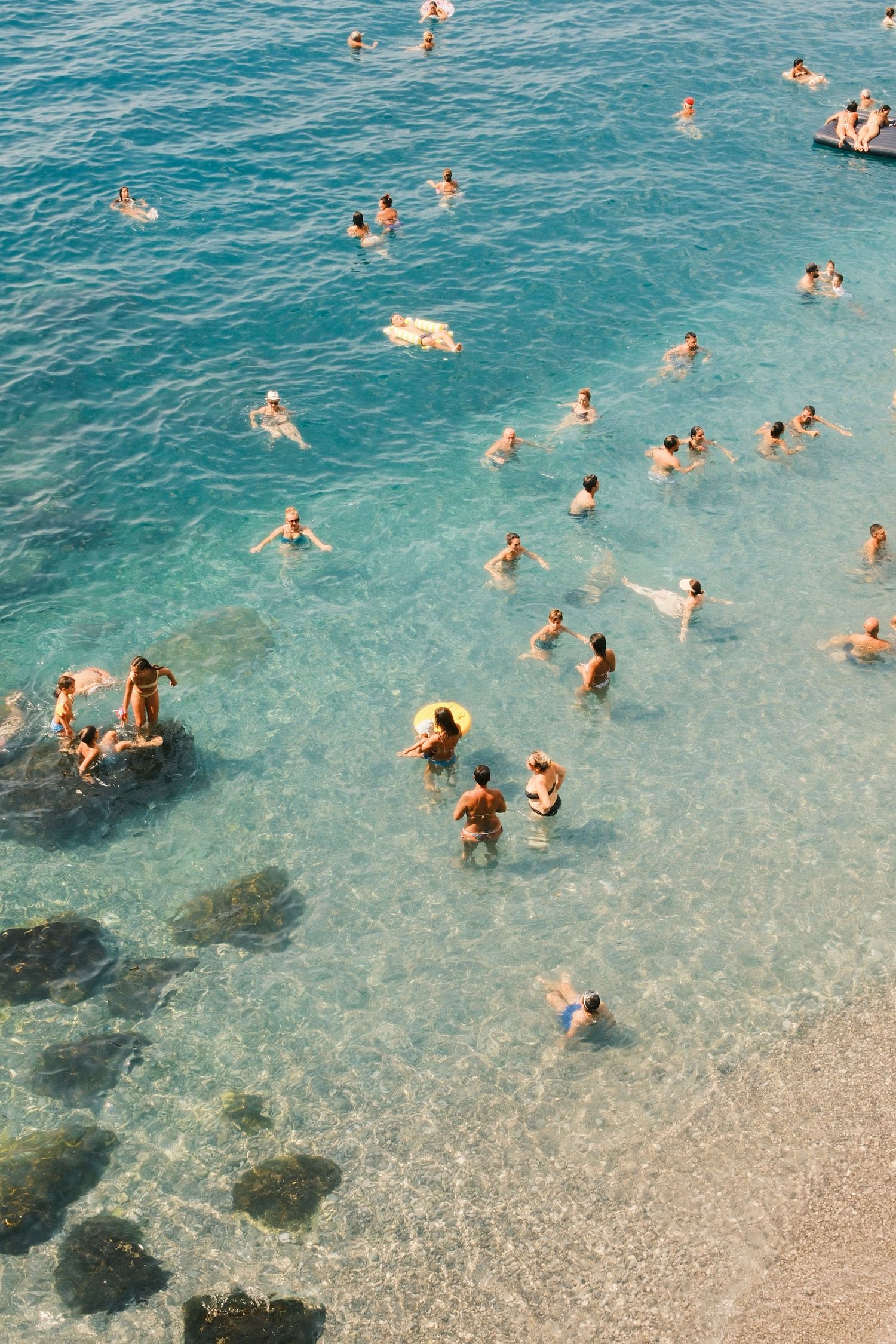 Aerial view of many swimmers enjoying crystal clear turquoise ocean water at a sunny beach resort.