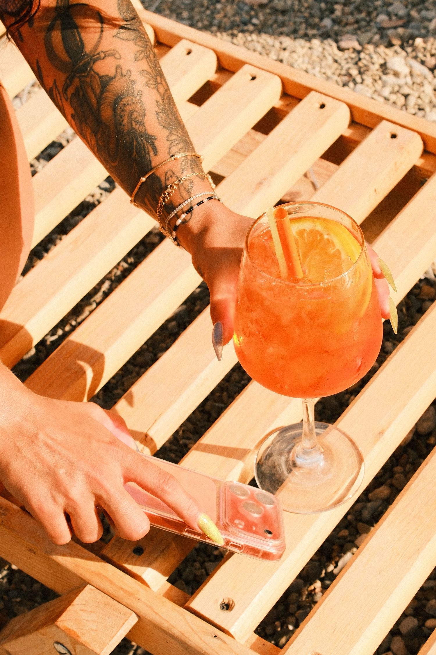 Tattooed arm reaching for a phone beside an Aperol Spritz cocktail on a wooden beach lounger.