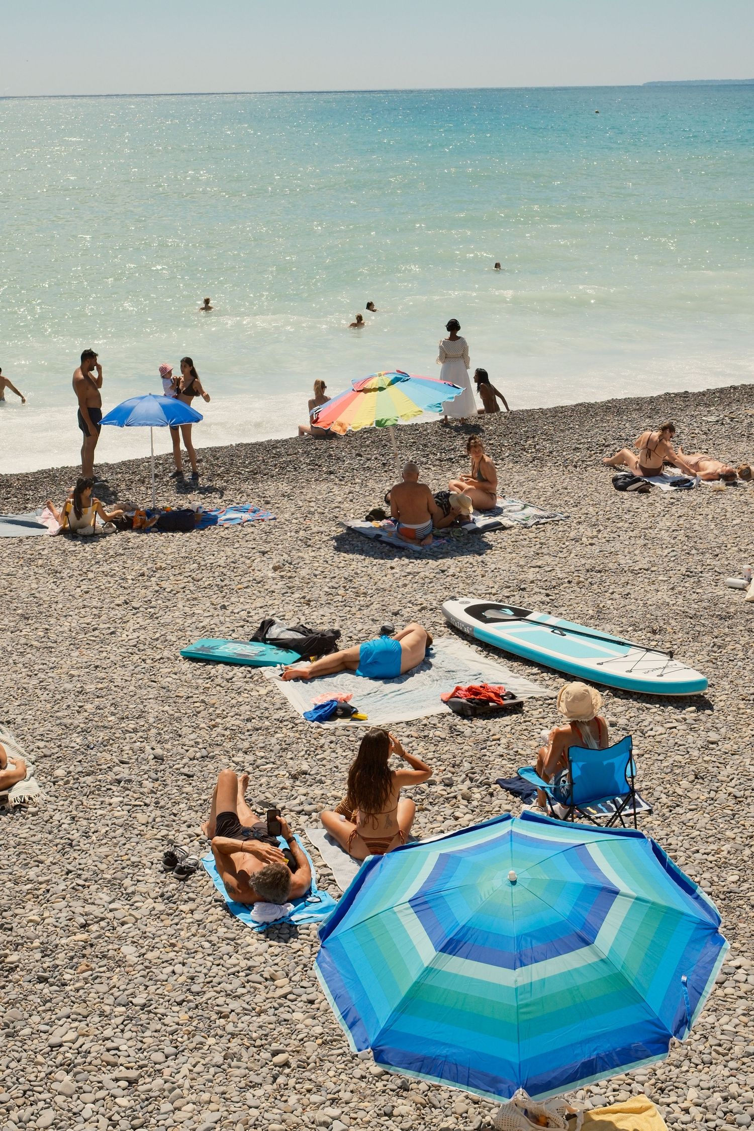Sunbathers relax on a pebble beach with colorful umbrellas, while swimmers enjoy the turquoise sea on a sunny day.